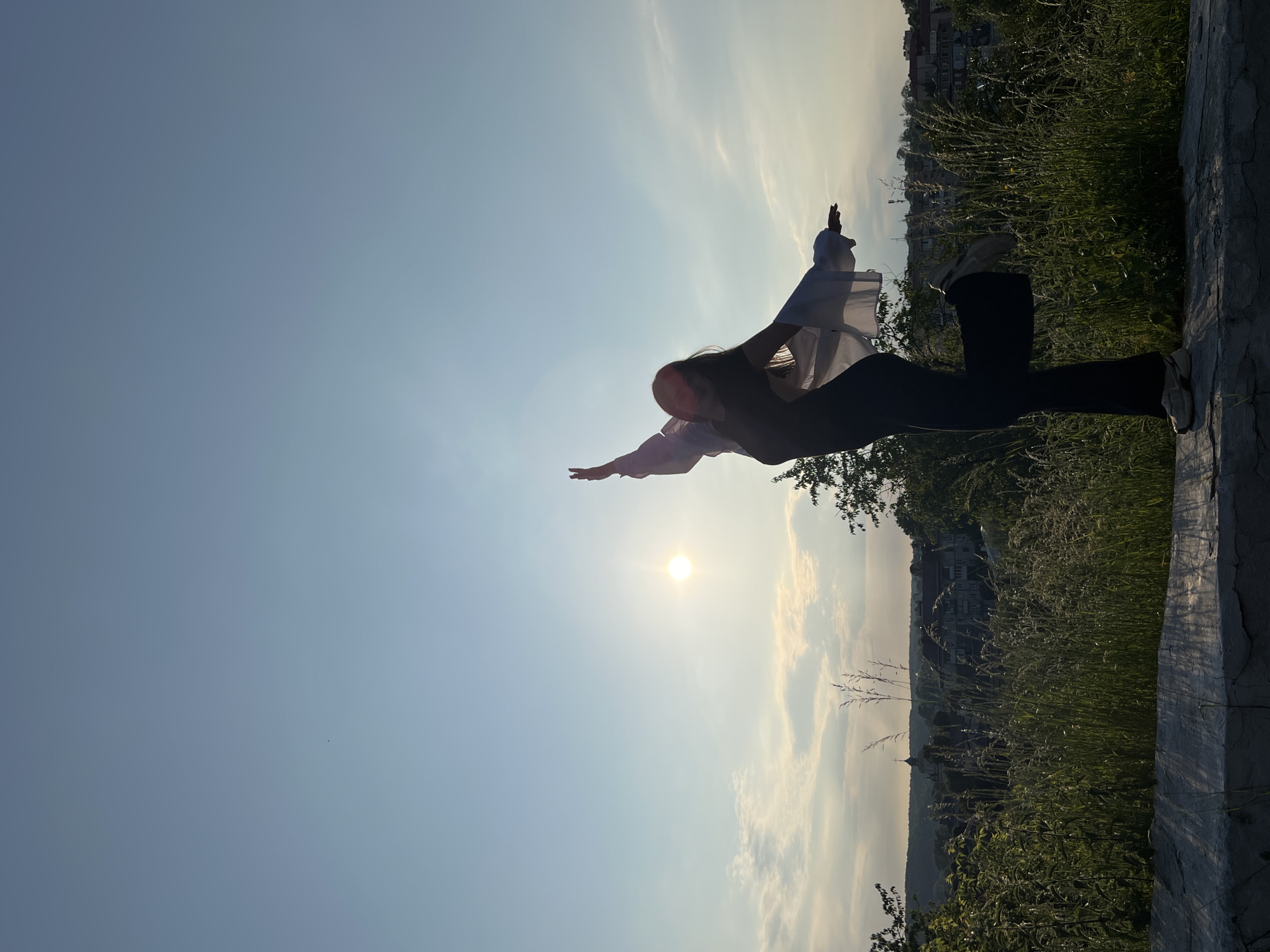 A full-body shot captures a person silhouetted against a bright, sunny sky, facing left with arms outstretched as if soaring or balancing. The person stands on a low concrete or stone wall, which forms the foreground and runs horizontally along the bottom-right of the frame. They are dressed in dark trousers, a dark top, and a light-colored, possibly sheer, long-sleeved outer garment that billows slightly around their arms.
The sun is visible in the mid-left of the clear blue sky, casting a strong backlight and creating a warm glow. Sparse, wispy clouds are scattered across the horizon, particularly on the right side of the frame. The background reveals a scenic landscape with rolling hills, abundant green vegetation, and distant buildings suggesting a town or city, consistent with Zbarazh, Ukraine. The person appears to be on an elevated vantage point overlooking the landscape.
The time of day appears to be late afternoon or early evening, indicated by the golden light and the sun's position, suggesting golden hour. The weather is clear and fair. The pose and setting evoke feelings of freedom, joy, and expansive tranquility. No visible text is present in the image.