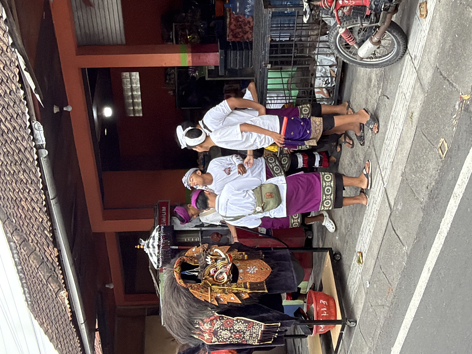 In North Kuta, Indonesia, a group of four young men stand on a sidewalk in front of a building with an orange facade and a tiled roof. The men are dressed in traditional Balinese attire, consisting of white shirts and purple sarongs. Three of the men are looking towards the left of the frame, where a large, elaborate costume, possibly for a Barong dance, is displayed. The costume is adorned with intricate gold and red patterns and topped with a decorative umbrella. One man, wearing a green shoulder bag, is facing away from the camera and appears to be speaking to the others. To the right of the group, a red motorcycle is parked. The sidewalk is paved with gray tiles and bordered by a white line, indicating it is a public street. The background shows a shop with shelves and various items for sale.