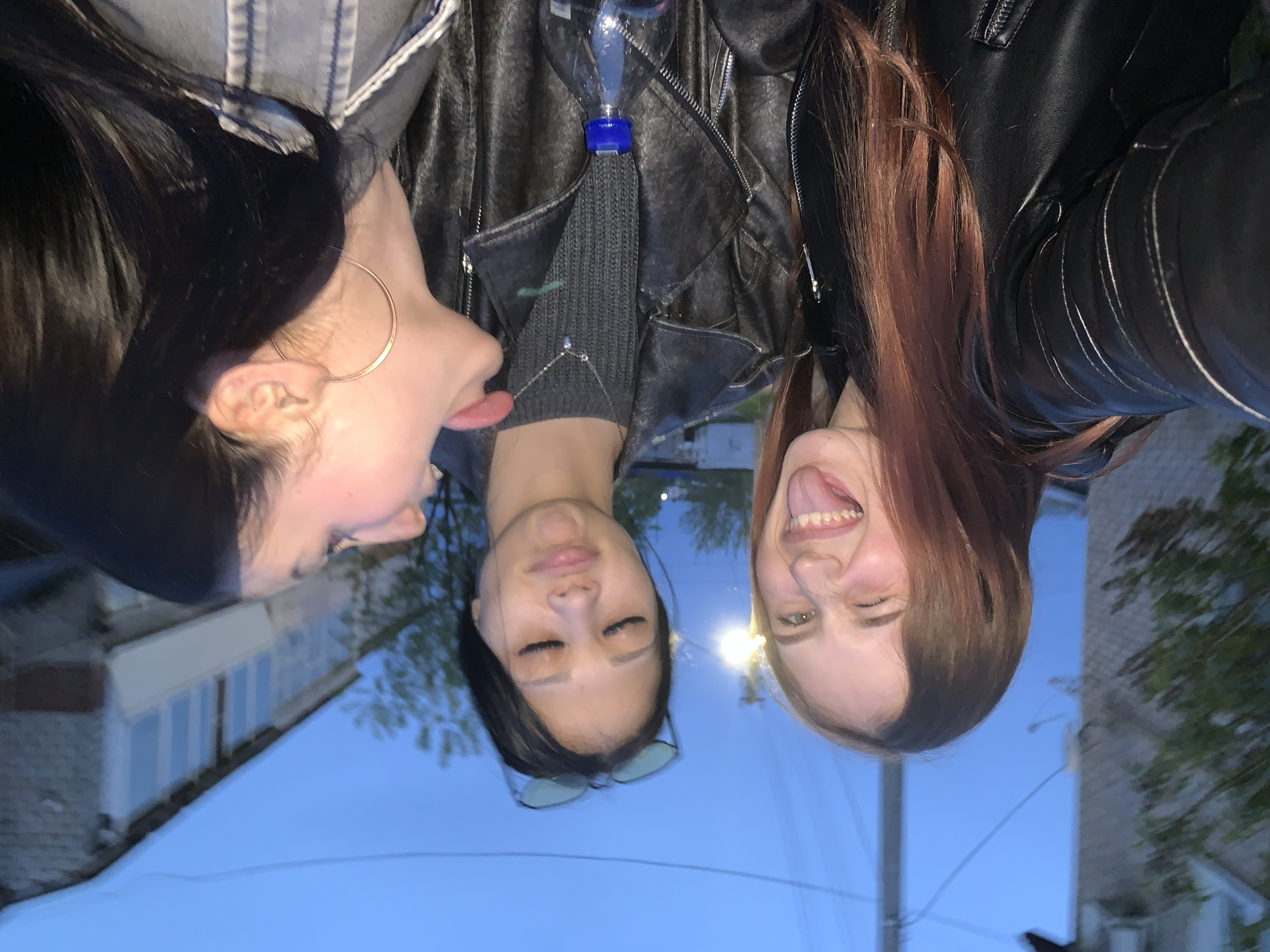 Three women are standing together outdoors in an urban environment at dusk. The woman on the left has long, dark hair and is wearing a large hoop earring, sticking her tongue out. The woman in the center has long, dark hair, closed eyes, and is wearing a dark gray shirt with a necklace and glasses. The woman on the right has long, reddish-brown hair and is sticking her tongue out. All three women are wearing dark jackets. A clear plastic water bottle hangs from a zipper of a leather jacket. In the background, buildings with lit windows, trees, and a street light can be seen, suggesting the scene is in Фоки, Russia.