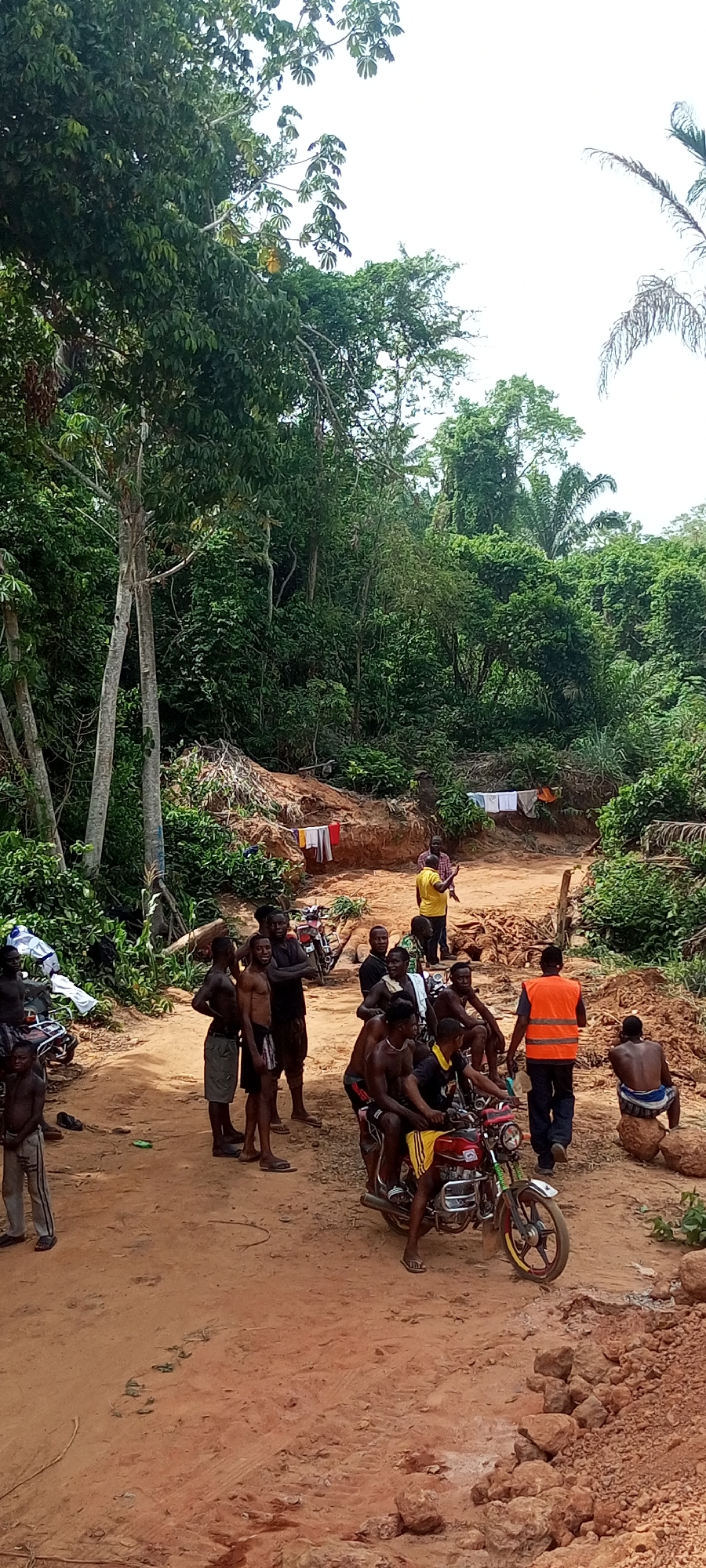 This media file captures a gathering of people, predominantly men, on a reddish-brown dirt road in a densely forested, tropical environment. The scene is set in Aloko, Nigeria, under bright daylight with a clear sky visible above the lush green canopy.
The main subjects are a group of individuals of varying ages, many shirtless or in simple attire, congregating on and around the unpaved road. Some stand casually, while others are seated, including two men on a red motorcycle with yellow detailing, and one man perched on a large rock. In the mid-ground, a man in a yellow shirt is actively gesturing or speaking to others, while further back, another man in a bright orange safety vest walks away. Two motorcycles are visible; the red one in the foreground and a less distinct one further back to the left.
The setting is characterized by thick, verdant jungle vegetation bordering the road, suggesting a rural or remote location. Mounds of reddish-brown earth flank the road, and clothes are seen drying on lines strung between trees or structures in the background, indicating human presence and daily life amidst nature. The overall atmosphere appears informal, possibly a communal gathering or a stop during travel. No visible text is present in the image.