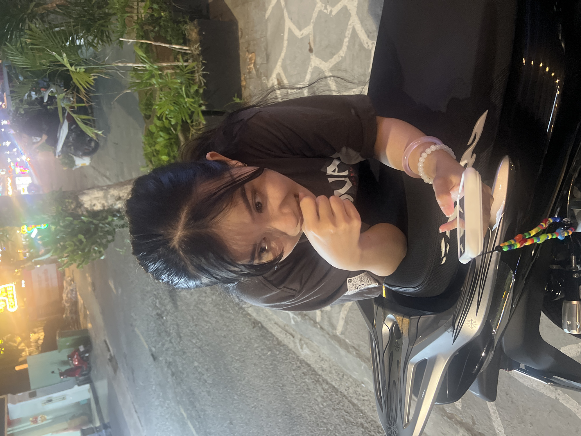 The image shows a young East Asian woman seated on a black and silver motorcycle or scooter at night, likely on a street or sidewalk in Đà Nẵng, Vietnam. She has long, dark hair and wears a dark t-shirt with a QR code printed on the right side of her chest. She is looking towards the camera with a slight smile, appearing relaxed.
In her right hand, she holds a white smartphone. On her right wrist, she wears two bracelets: one light purple and another composed of white beads. Her left hand is brought up towards her chin or mouth. The motorcycle she is sitting on features distinctive silver and black accents with snowflake-like or abstract patterns. A colorful beaded charm is attached to the motorcycle, near the handlebar area.
The background indicates nighttime, with a dark sky and illuminated elements. Bright, colorful artificial lights, possibly from establishments or street vendors, are visible on the left side of the frame, casting a glow. Green foliage, including palm-like plants, is present behind her. The ground is a paved surface, possibly a sidewalk, with light-colored patterns. In the far background, a light blue-green building and another red scooter are faintly visible, contributing to the urban street setting. No discernible text is visible apart from the QR code on her shirt.
