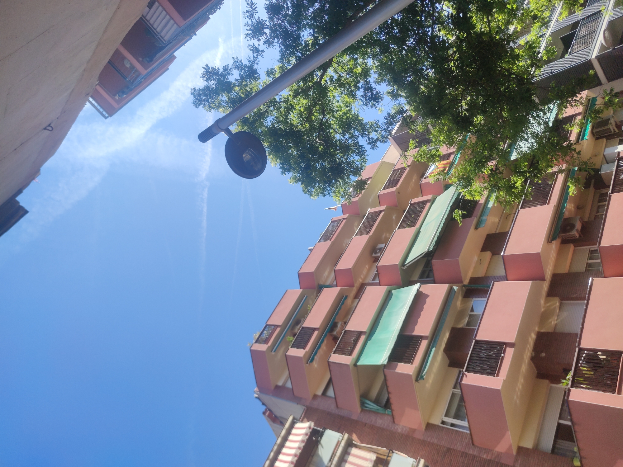 The image, taken from a low angle looking upwards, features a scene in the city of Unknown, Unknown. The primary subject is a tall building with a distinctive architectural design. The building's facade consists of alternating colors, primarily a reddish-brown and a lighter beige, with cuboid extensions that appear to be balconies or enclosed spaces. Some balconies have green awnings extended, providing shade.
A street lamp, with a dark, round shade, is visible in the upper left of the frame. A tree with green foliage is positioned to the right of the lamp, its branches partially obscuring the upper portion of the building.
The sky, a clear blue, forms the background for the composition. Contrails, or condensation trails from airplanes, can be seen streaking across the sky. Another building, partially visible, is in the upper left corner of the frame.