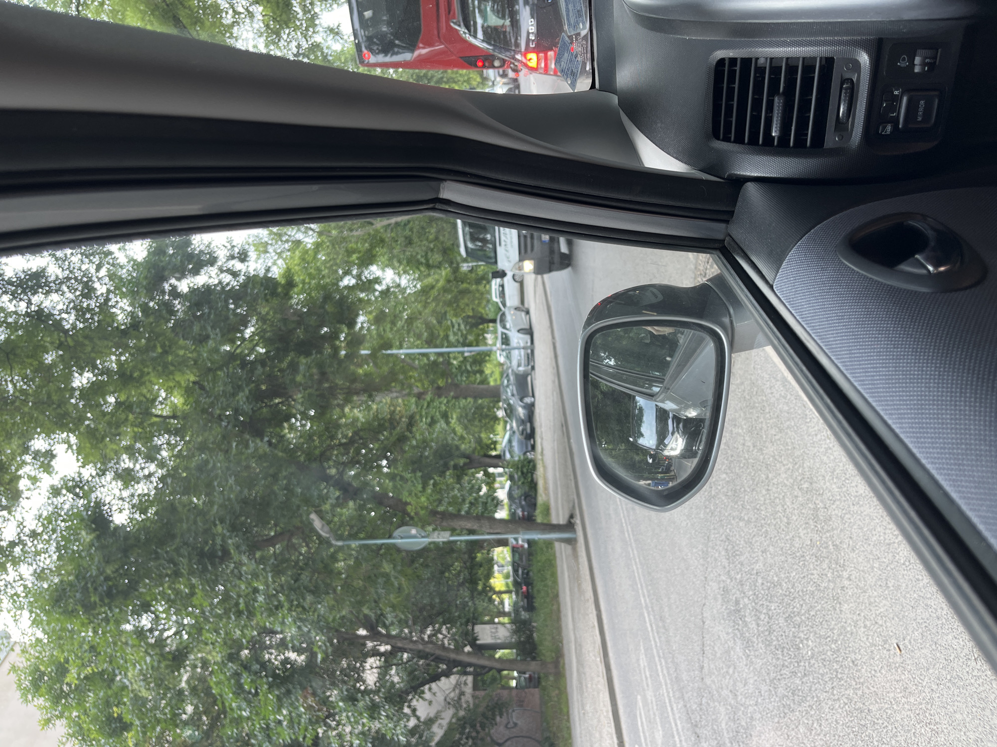 The image captures a view from inside a car, looking out the passenger side window and reflecting in the side mirror. The car is in traffic on a street lined with trees. Several other vehicles are visible, including a white van, a red bus, and various cars. The street appears to be paved and has lane markings. In the background, there are buildings and more trees. The side mirror reflects a black car and pedestrians on the sidewalk. The interior of the car is also partially visible, showing part of the dashboard and door panel. The scene is consistent with a street in the city of Orizari, Bulgaria, given the context provided.