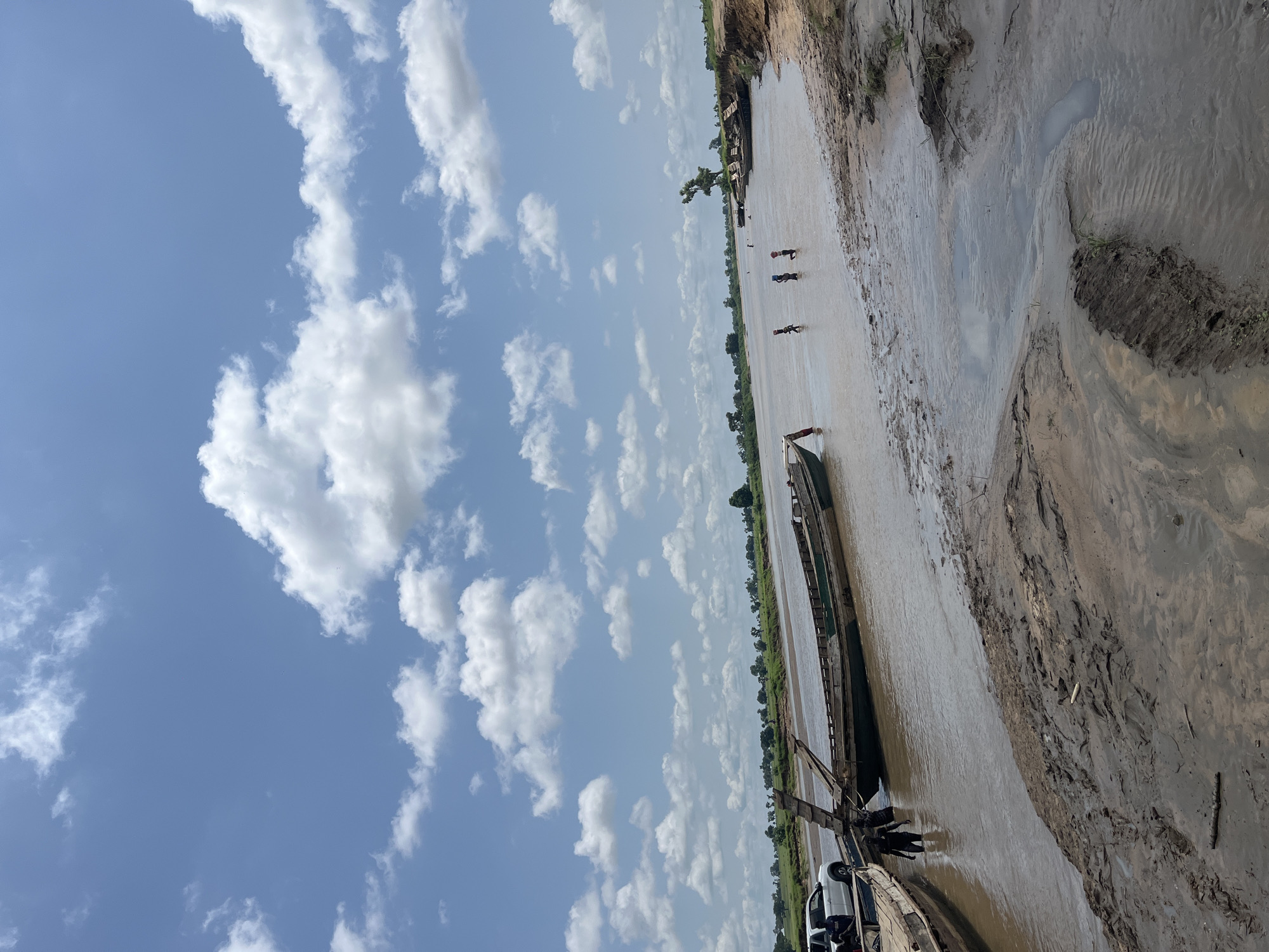 The image depicts a wide, muddy river with several wooden boats docked along the bank. The water is a murky brown, and the riverbed appears to be composed of mud and sand. In the foreground, the muddy bank is uneven with some vegetation growing out of it.
There are several boats in the water, some are partially submerged or grounded on the mud. On the left side of the image, a white vehicle, possibly a pickup truck, is parked near the boats, and two individuals are visible near the water's edge. Further into the river, in the middle ground, a group of at least three children are wading in the water. In the background, the opposite bank of the river is lined with green vegetation and scattered trees, suggesting a rural or semi-rural environment. The sky is blue with scattered white cumulus clouds.
The scene appears to be a riverbank in a natural setting, possibly a fishing or transportation hub. The presence of boats and people engaged in activities in the water indicates this is likely a functional location for the local community. The general atmosphere is bright and sunny, suggesting daytime. The specific location provided is Nemnei, Nigeria.