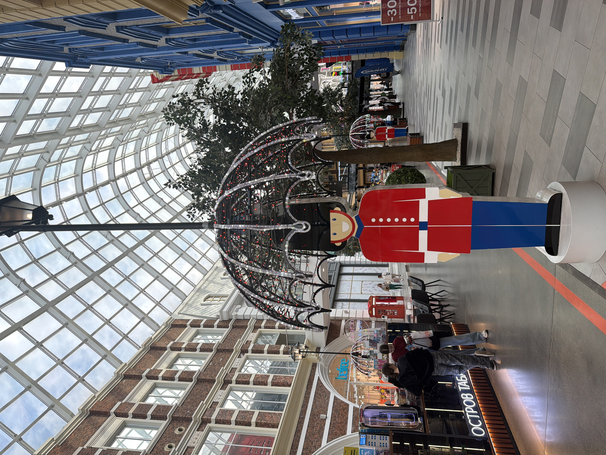 The image depicts a well-lit indoor shopping mall with a distinctive glass ceiling. The architecture of the mall appears to be themed, with buildings on either side of the walkway designed to resemble traditional brick and blue facade structures. Dominating the foreground is a large, decorative statue of a British royal guard, holding an illuminated umbrella. The guard is dressed in a red tunic with white accents and a black bearskin hat, with blue trousers. In front of the guard, a red line is marked on the grey tiled floor.
To the left of the guard, several people are interacting at a counter with neon lighting that displays Russian text. There is also a red British-style telephone booth visible. A large artificial tree is placed behind the guard statue. Further down the walkway, other decorative elements, including smaller versions of the guard statue with umbrellas, are visible, along with more trees and shop facades. People are seen walking and browsing in the distance. The overall impression is a festive and decorative commercial environment.