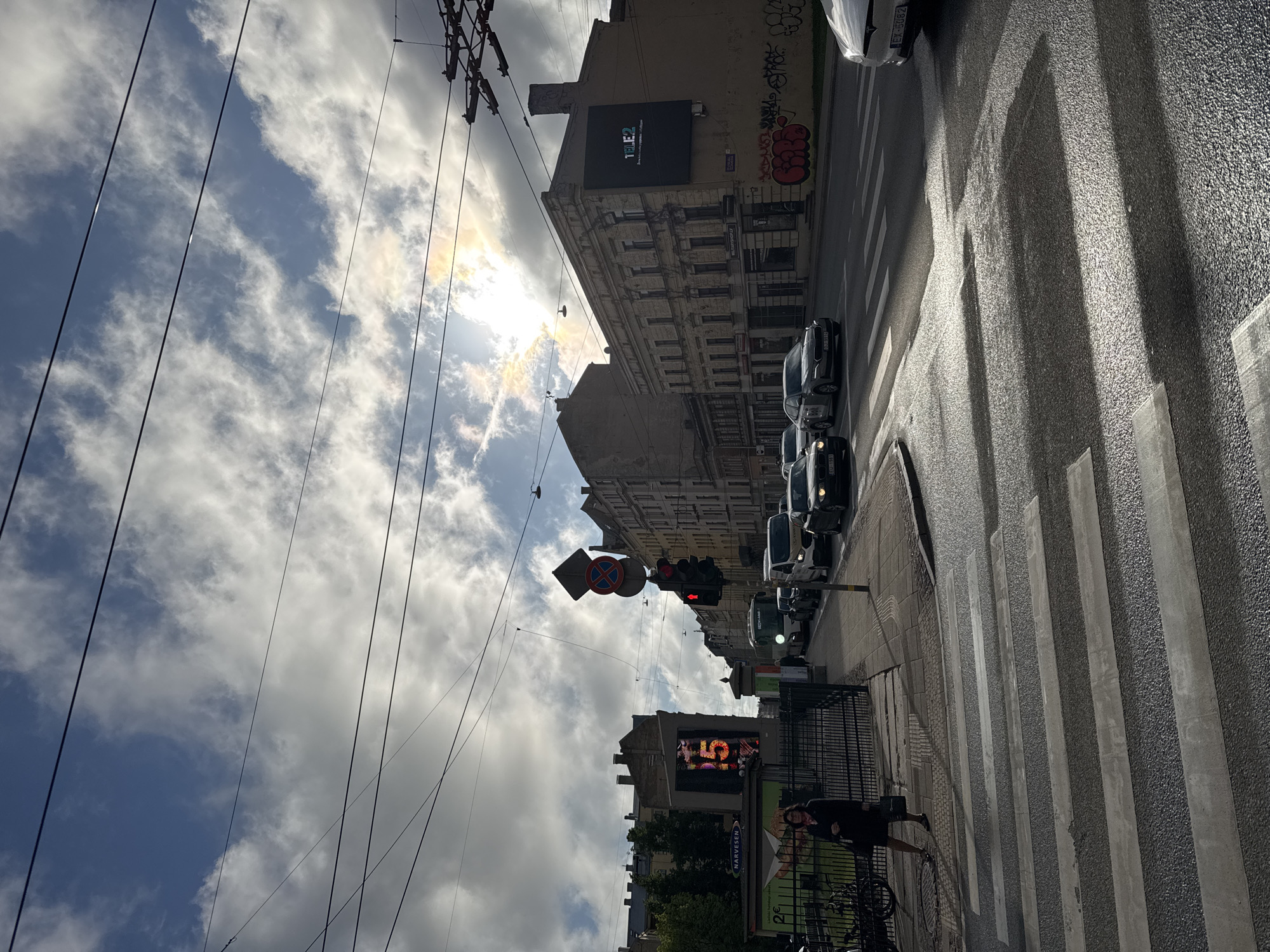 The image depicts a street scene in Riga, Latvia, viewed from a ground-level perspective, rotated counter-clockwise by 90 degrees.
Sky: The sky is partially overcast with patches of blue visible amidst the clouds. The sun appears diffused behind the clouds, creating a bright area in the sky. Electrical wires are stretched across the sky in the foreground.
Buildings: Buildings line the street. The facades are of varying styles and architectural periods. One building features a large black digital billboard on its upper portion, displaying the text "TELE2". Graffiti is visible on the side of one of the buildings. Another building features a digital advertisement on the lower corner, next to a Narvesen advertising poster.
Street: The street is wet and reflective, indicating recent precipitation. A zebra crossing is visible, with clear alternating white and gray stripes. Multiple cars are lined up, stopped at a traffic light that is currently red. There is one pedestrian visible walking along the sidewalk.
Traffic Signals: A traffic signal is visible, displaying a red light. A 'no entry' road sign is located above the traffic signal.
People: One pedestrian, dressed in black, is visible walking on the sidewalk, near a bicycle parking area.