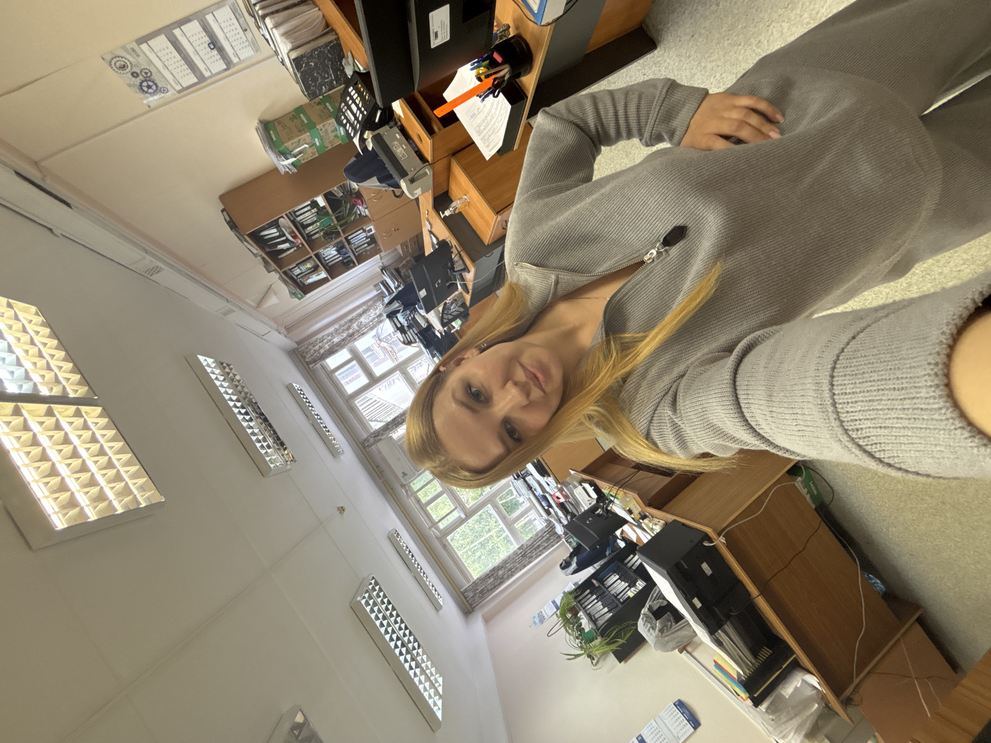 The image shows a selfie taken in what appears to be an office. A woman with blonde hair is in the foreground, wearing a gray zip-up sweater or jacket. In the background, there are office desks, cabinets, and what seems to be filing cabinets. Multiple windows provide natural light, and there are fluorescent lights in the ceiling. There are office supplies and equipment on the desks. No information about location being in the city of Rusanovo, Russia can be conclusively determined from the image alone.