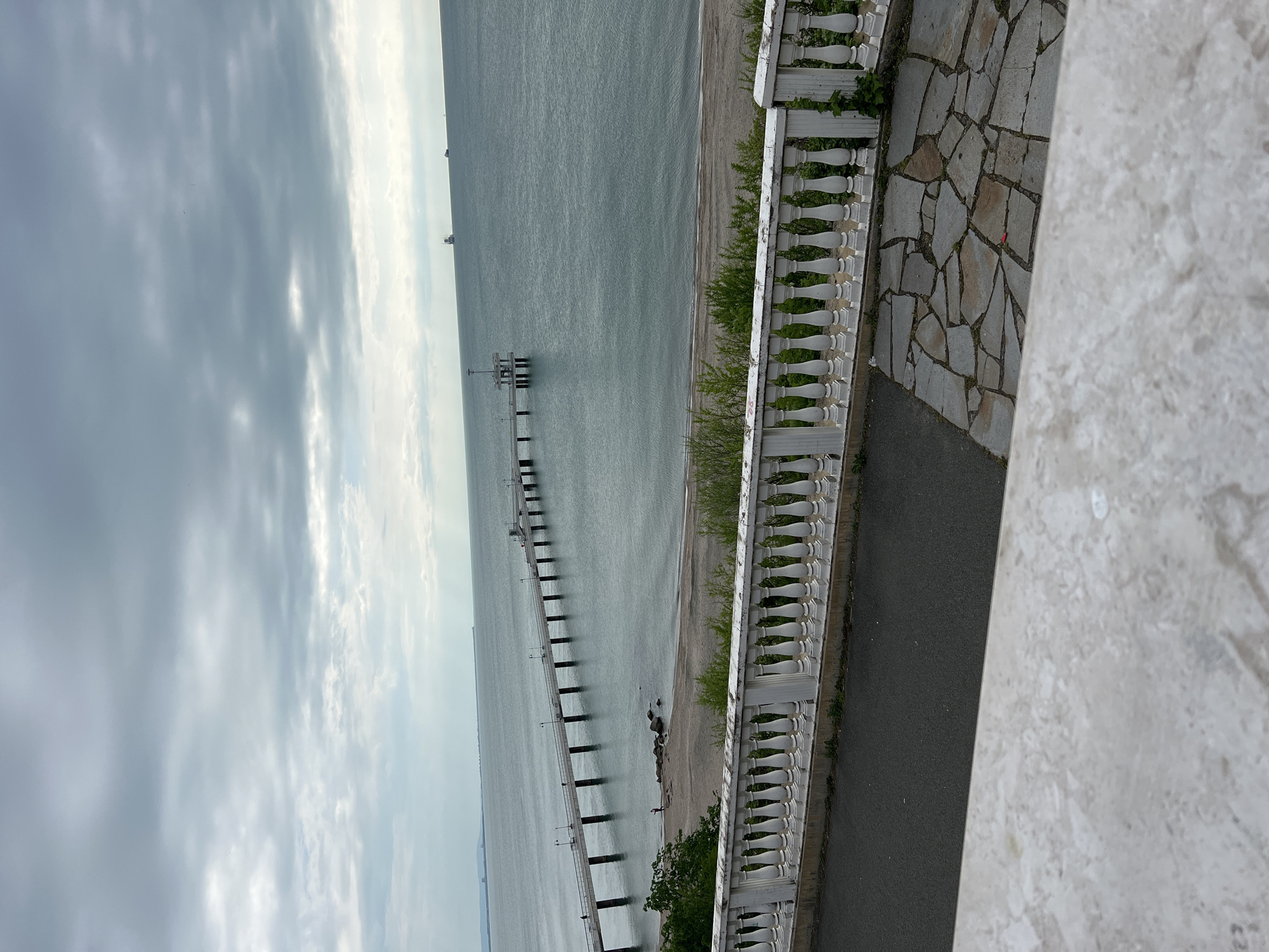 The image depicts a view of Burgas Bridge in Burgas, Bulgaria, from an elevated position. A long pier extends into the sea under a cloudy sky. The right side of the frame features a white balustrade, a cobblestone walkway, and a glimpse of a gray asphalt surface.