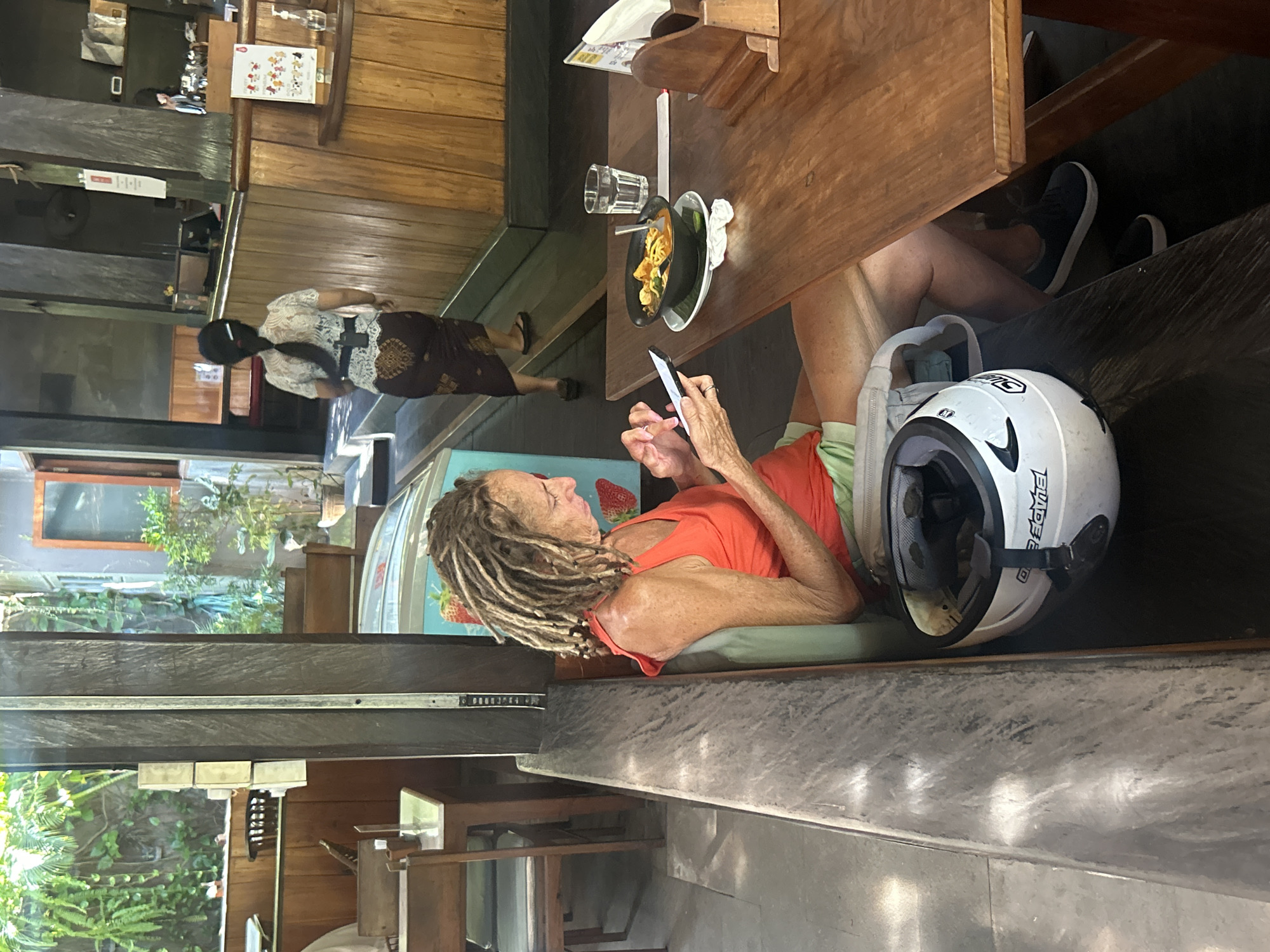 In North Kuta, Indonesia, a woman with dreadlocks, wearing an orange tank top and light green shorts, sits on a bench at an outdoor seating area of a restaurant. She is using her smartphone, and a white helmet with black accents is on the bench next to her. On a wooden table in front of her, there is a bowl of food, a glass of water, and a napkin holder. In the background, a person wearing traditional Balinese attire is walking away from the camera. The environment appears to be a tropical setting with lush greenery visible through an open doorway.