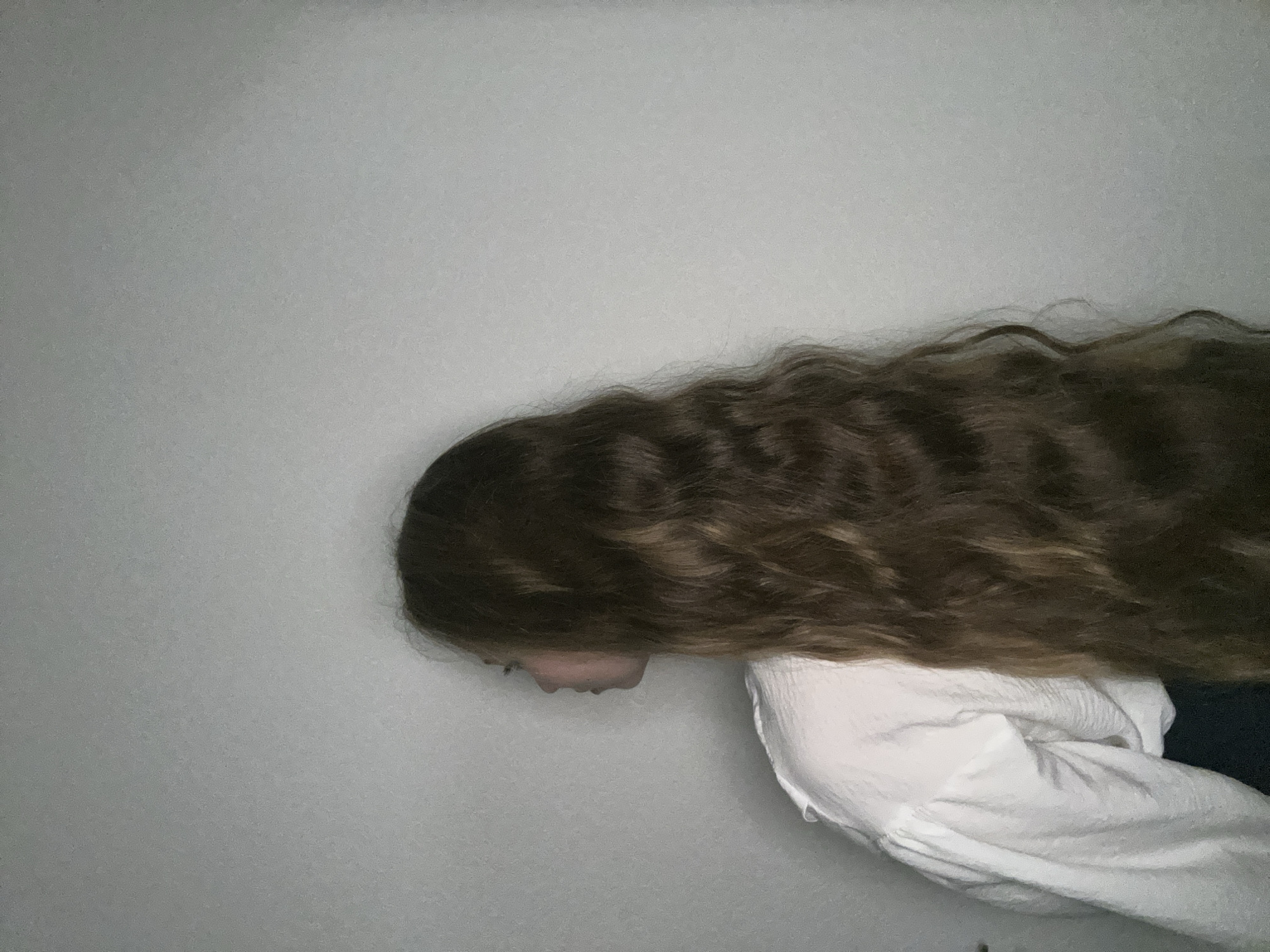 A person with long, wavy, brown hair and wearing a white shirt is leaning forward against a light gray wall. The person's head is angled downwards, with only a portion of their face visible in profile. The image appears to be taken indoors, with the wall serving as the background. There are no indications of Saint Petersburg, Russia, in the image.