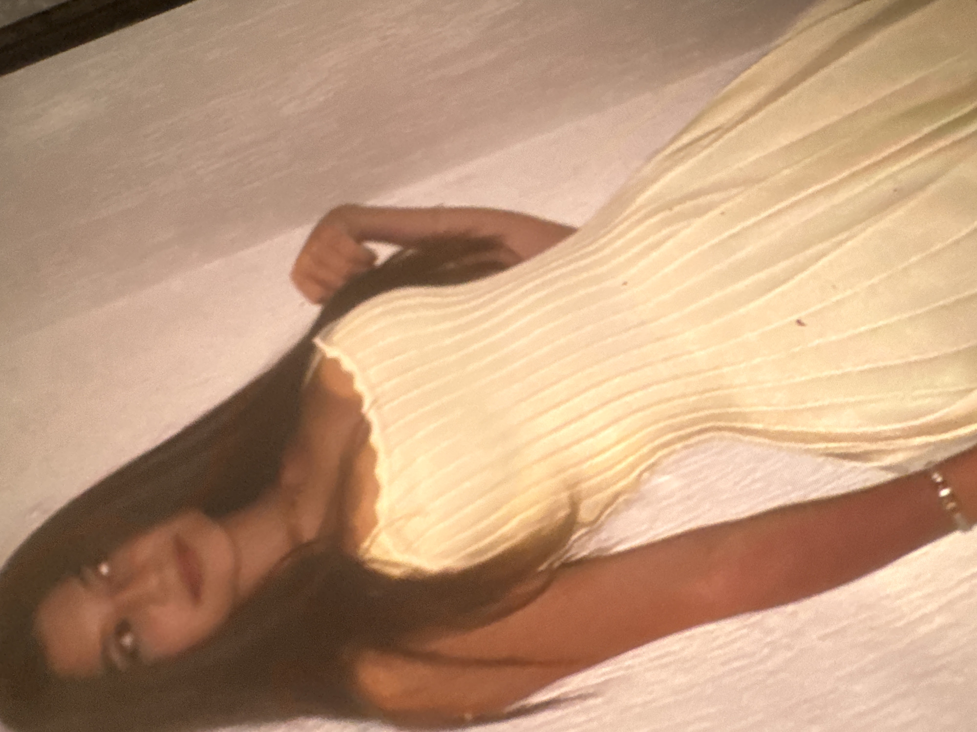 A young woman with long, dark hair is depicted lying down indoors, angled diagonally across the frame. She is wearing a light-colored, possibly cream or light yellow, pleated garment, which appears to be a top or dress. Her face is turned towards the viewer, though it is slightly out of focus and blurred. Her expression appears calm or neutral. A faint glint on her right wrist suggests she might be wearing a bracelet or watch. The background is a plain, light-colored surface, possibly a bed or floor, with a dark horizontal line visible in the upper left corner, potentially a piece of furniture or shadow. The overall lighting is soft, giving the image a somewhat warm and relaxed tone. The image quality is a bit grainy and blurred, suggesting it may have been taken in low light conditions or with an older camera. There is no visible text in the image.