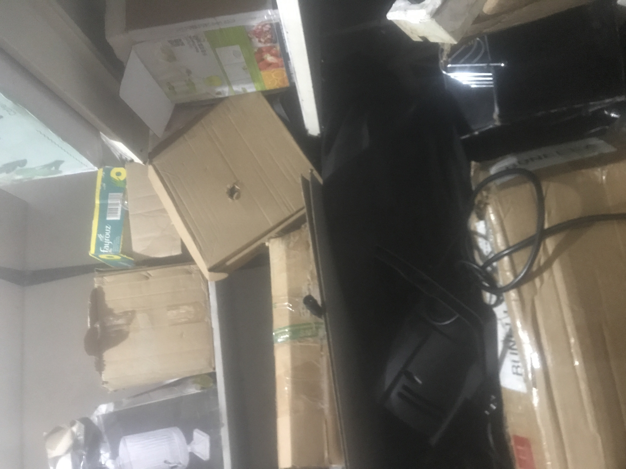 The image displays a cluttered indoor storage area, appearing to be a room or corner filled with various items. The perspective is tilted, with the scene dominated by numerous cardboard boxes of different sizes and conditions, stacked and placed somewhat haphazardly. Many boxes appear worn; one large cardboard box in the foreground exhibits a prominent dark, irregular stain.
Among the visible objects is a distinctive green and yellow box labeled "Fayrouz." Another white box features product imagery, including fruits like strawberries and oranges, suggesting it might contain a food or kitchen-related item. A large, dark-colored electronic device, possibly a monitor or television, is prominently positioned towards the right side of the frame, with black electrical cables intricately coiled around it and draped over other boxes. In the lower left section, a white, ribbed cylindrical object, resembling a fan or an air purifier, is partially visible. Several packages are wrapped in clear plastic tape.
No people are visible, and no specific activity is taking place; the scene depicts a static arrangement of stored goods. The lighting is somewhat dim and uneven, creating a subdued atmosphere. Visible text includes "Fayrouz" on a green and yellow box and a partial word, "BUM," on a brown cardboard box in the lower right area. The overall impression is one of a domestic or commercial storage space.
