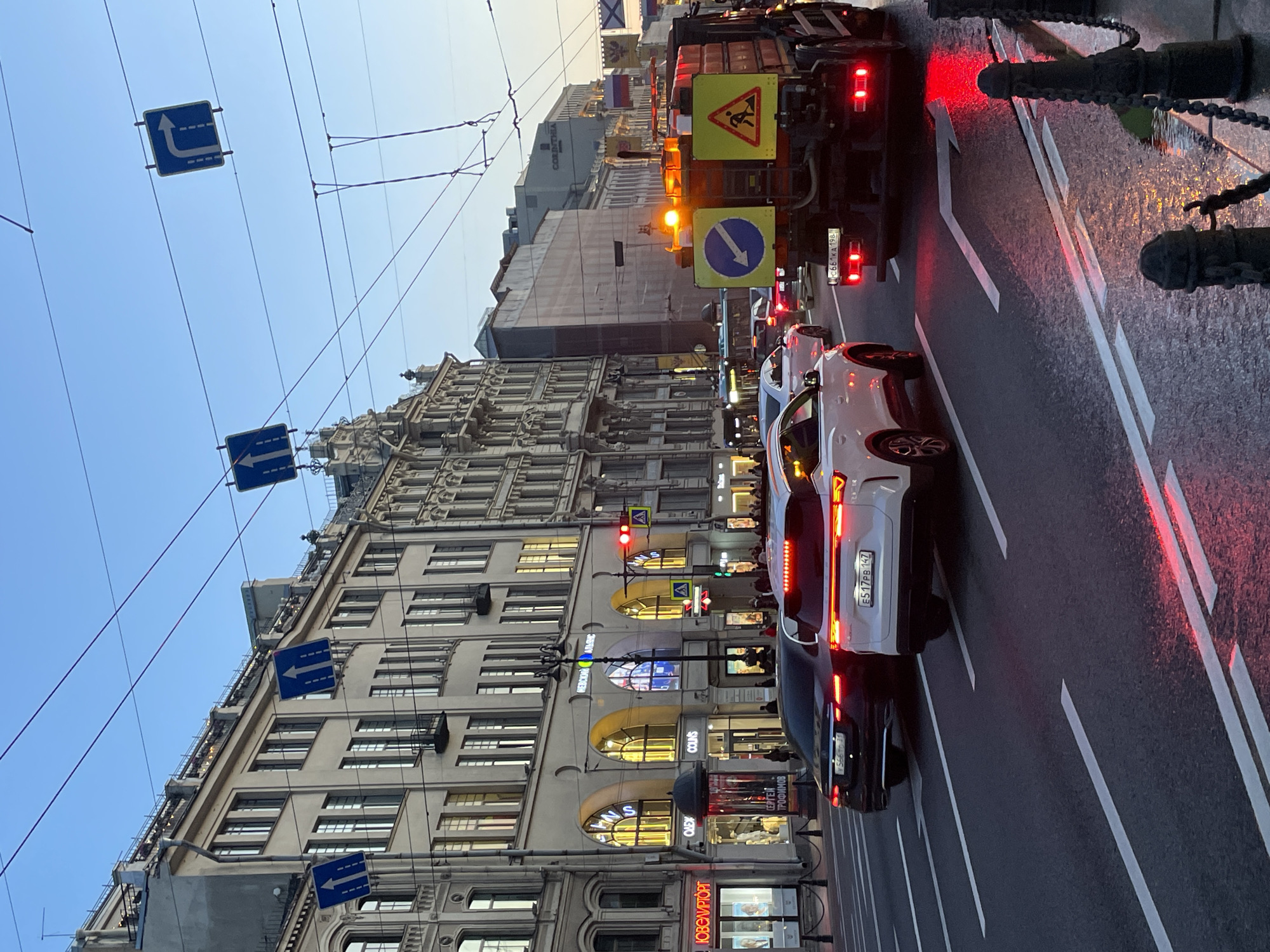 The image captures a bustling street scene in Saint Petersburg, Russia, likely during dusk or early evening, indicated by the fading light in the sky and illuminated shop windows. The perspective is looking down a wide urban street, with traffic in the foreground and ornate buildings lining the left side.
In the foreground, a large utility truck with amber warning lights and two yellow warning signs is stopped in traffic. One triangular sign depicts a person with a shovel, indicating roadwork, and another circular blue sign with a white arrow points downwards to the right, suggesting a direction to pass. The truck's red brake lights are illuminated. Behind the truck, a white car (likely an SUV) and a darker sedan are also stopped, their red brake lights glowing. License plates "E517PB 47" (white car) and "C661AA 198" (truck) are clearly visible, confirming the Russian location (region codes 47 and 198 are for Leningrad Oblast and Moscow, respectively, though the scene is in Saint Petersburg itself). The asphalt road appears wet and reflective, possibly from recent rain.
To the left, a row of grand, multi-story buildings with classical architecture features numerous windows and decorative elements. The ground floors house various shops and businesses, with arched windows and glowing interior lights. Visible shop names include "СКОРНЯК" (Skornyak, meaning furrier), "COLLINS," "ОВСОВТОРГ" (OvsovTorg), and "СЕРГЕЙ ПРОФИМОВ" (Sergey Profimov). A red traffic light is visible on the sidewalk. Overhead, a network of wires, likely for trolleybuses or trams, crisscrosses the sky. Several blue square road signs with white arrows, indicating lane directions (e.g., straight, turn left), are suspended from these wires. Further down the street, a Russian flag and a naval flag (white with a blue St. Andrew's cross) are seen on building facades. The overall impression is that of a busy urban environment at the transition from day to night.