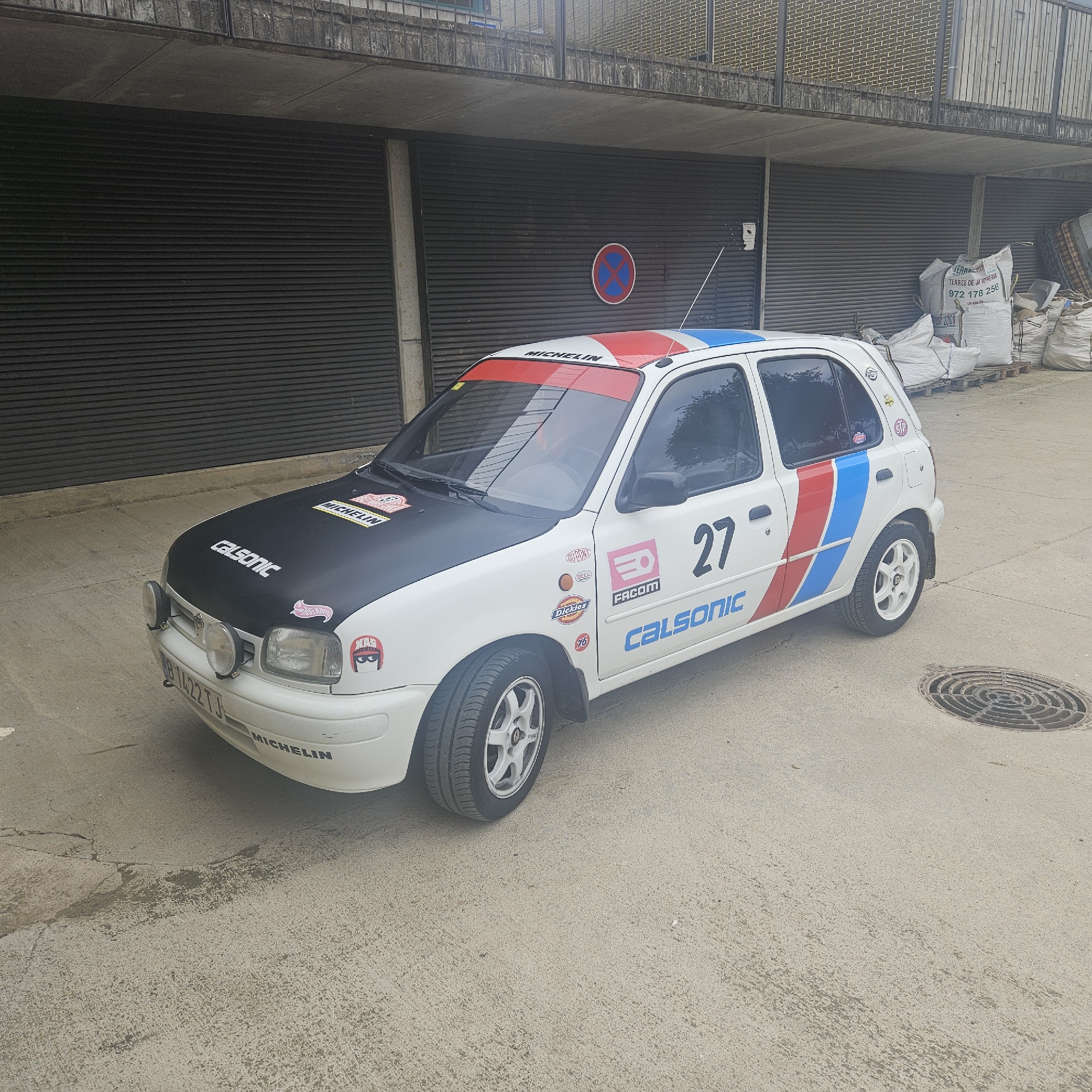 The image shows a white Nissan Micra rally car parked on a concrete surface in front of a dark, corrugated metal garage door. The car has a black hood with racing decals, including "MICHELIN" and "Calsonic". The sides of the car are decorated with red, white, and blue stripes, the number "27", and various sponsor logos such as "FACOM", "Dickies", and "STP". The car also has auxiliary headlights on the front bumper. In the background, there are several large white bags, possibly containing construction or gardening materials, stacked on pallets. Above the garage door, there is a concrete overhang with a metal railing. A circular manhole cover is visible on the ground to the right of the car. The location is identified as Aiguaviva, Spain.