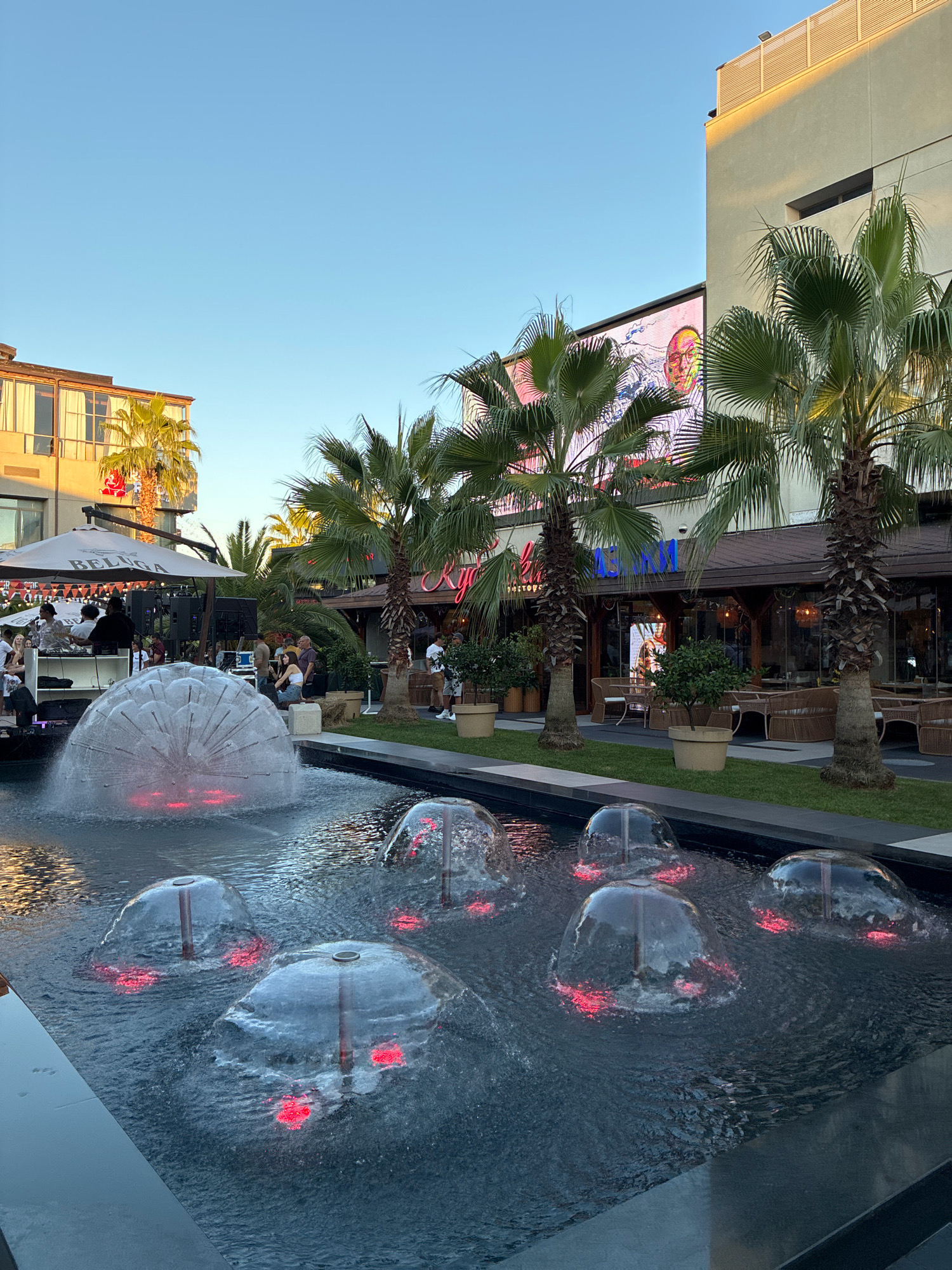 The image depicts an outdoor scene at what appears to be a commercial or entertainment district with a contemporary ambiance. In the foreground, a water fountain features several transparent, dome-shaped water jets with red LED lights illuminating them from below. The fountain is set within a dark-tiled pool.
Behind the fountain, there is a well-manicured lawn and a paved walkway. Further back, a group of people are gathered near a DJ booth under a large white umbrella. Some individuals are seated, while others are standing, and it seems like a social event or performance is taking place.
Dominating the background are modern buildings with several levels. Palm trees are scattered throughout the scene, suggesting a warm climate or a designed landscape. One building prominently displays a large digital screen showing an image of a man's face. Restaurants and seating areas are visible within the buildings, indicating a commercial establishment. The overall lighting suggests it is late afternoon or early evening, with a clear blue sky overhead. The specific location of Sochi, Russia, is not ascertainable from the visual elements in the image alone.