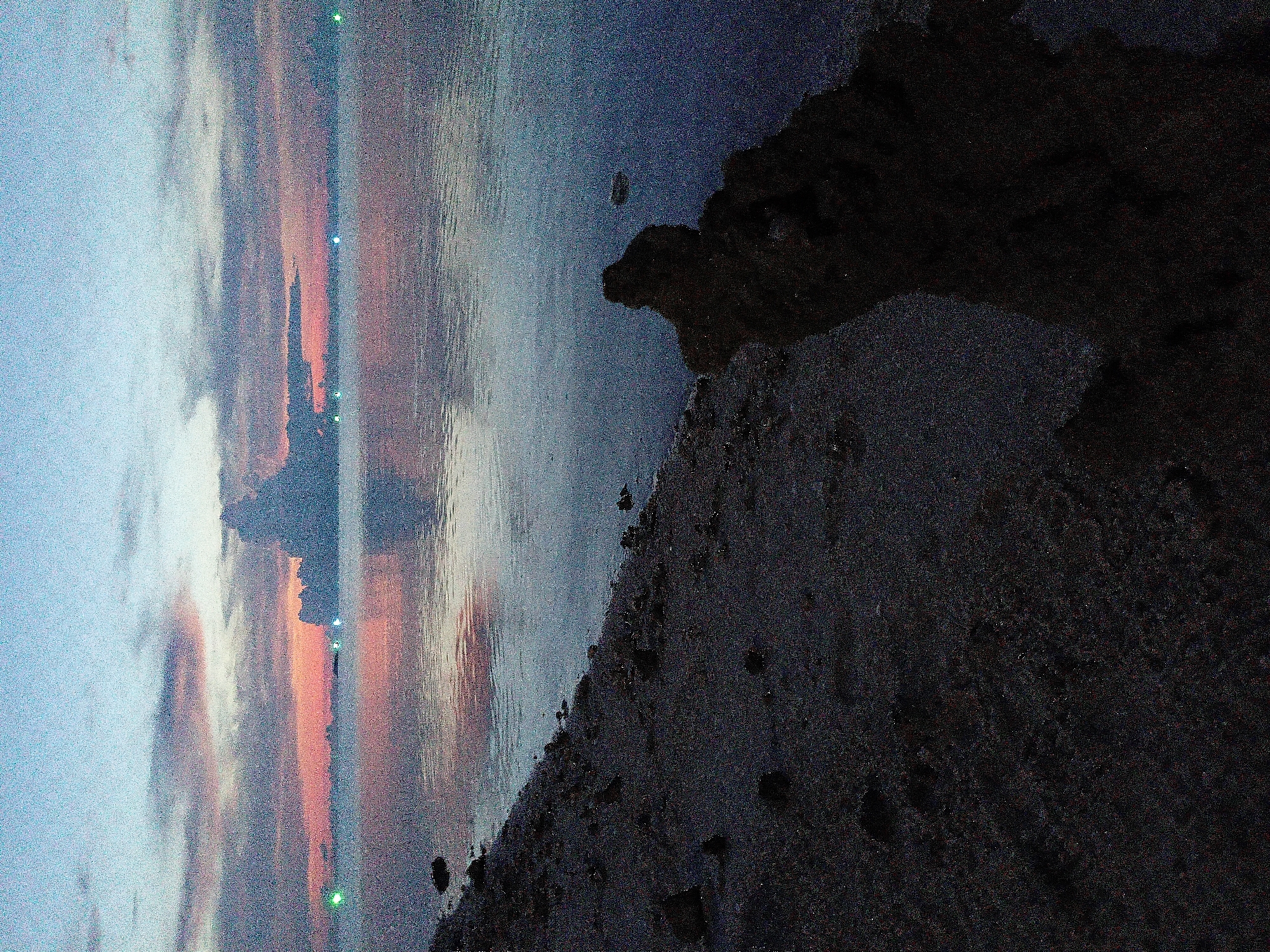 The image depicts a coastal scene at dusk in Baan Wok Tum, Thailand. The view is oriented with the shoreline on the right side of the frame. A dark, rocky landmass extends into the frame from the lower right, with a beach composed of dark sand and rocks. The water occupies the middle ground of the image, with a smooth surface reflecting the sky.
The sky shows a transition from light blue near the top to a mix of orange and pink hues towards the horizon, which is positioned to the left of the frame. Several small green lights are visible along the horizon. Clouds are present, reflecting the colors of the sunset. No people or other distinct objects are visible besides the rocks, sand, water, and sky.