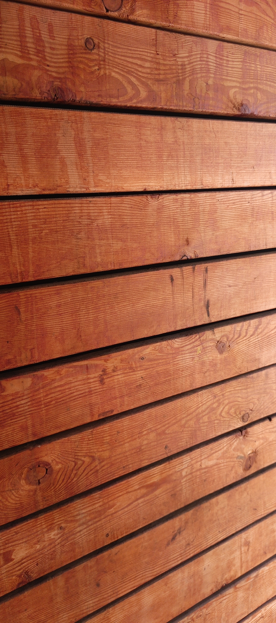 This image is a close-up, angled shot of a weathered wooden wall. The wood planks are oriented horizontally and are a warm, reddish-brown color, likely due to staining or age. The grain of the wood is prominent, with visible rings and occasional knots. There are dark, narrow gaps between the planks, creating a striped effect. The surface of the wood appears slightly textured and shows some signs of wear, with minor imperfections and smudges. The lighting suggests it is daytime, with shadows cast by the gaps between the planks.
There is no information available to associate this image with the city of Gorno-Altaysk, Russia.