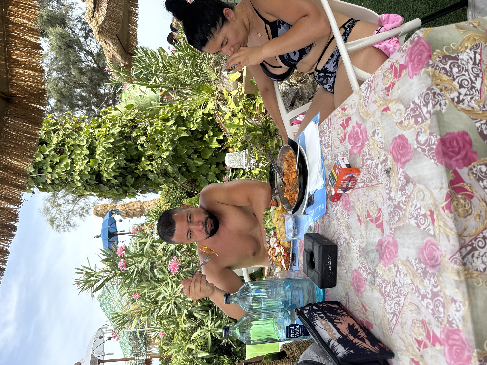 A shirtless man with a beard is eating food at a table on a patio in Cales De Mallorca, Spain. He is holding a fork with food on it and looking at the camera. A woman in a bikini is sitting to his right, drinking from a glass. The table is covered with a floral tablecloth and has several items on it, including bottles of water, plates of food, a cigarette pack, an ashtray, and a purse. In the background, there are palm trees, flowers, and umbrellas. The sky is partly cloudy.