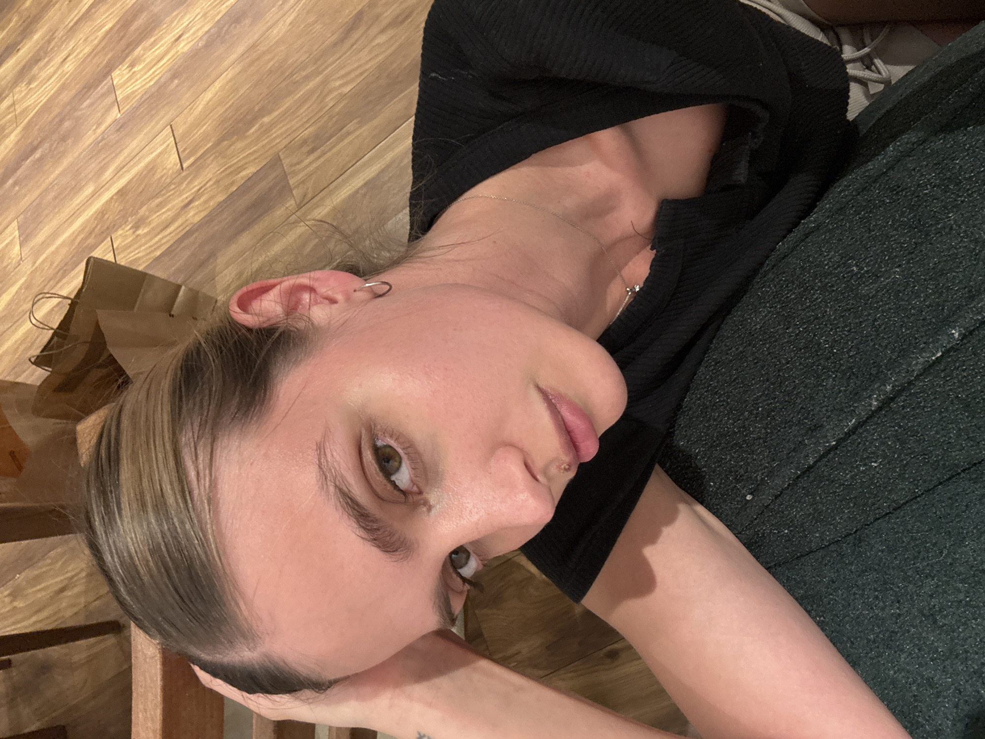 A person lies on a wooden floor, their head resting on their arm. They wear a black top and dark green trousers. A shopping bag sits near their head. The location is Unknown, Unknown.