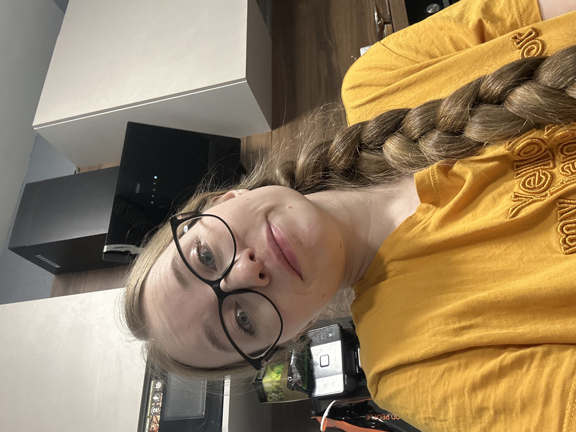 A young woman is depicted in an indoor setting, likely a kitchen or common room, with the image rotated sideways. She has light brown hair styled in a thick, long braid draped over her left shoulder and chest. She wears black-rimmed glasses and a mustard yellow long-sleeved shirt with embroidered text on the front, partially visible as "OR" and "OOD PEOPLE." Her eyes are light-colored, and she has a subtle, closed-mouth smile, looking directly towards the camera.
In the background, to her left, a black Haier brand range hood with a small blue indicator light is visible above a dark surface. Above the range hood are white upper cabinets. To her right, a black electronic device, possibly a smartphone or tablet, rests near a green and white package and another item with "HYUNDAI" text. Further back, dark wooden cabinetry or wall paneling is visible. The overall lighting is bright and even, typical of indoor artificial light. The scene suggests a casual moment, possibly a selfie or a relaxed pose, in what appears to be a modern home in Saint Petersburg, Russia.
