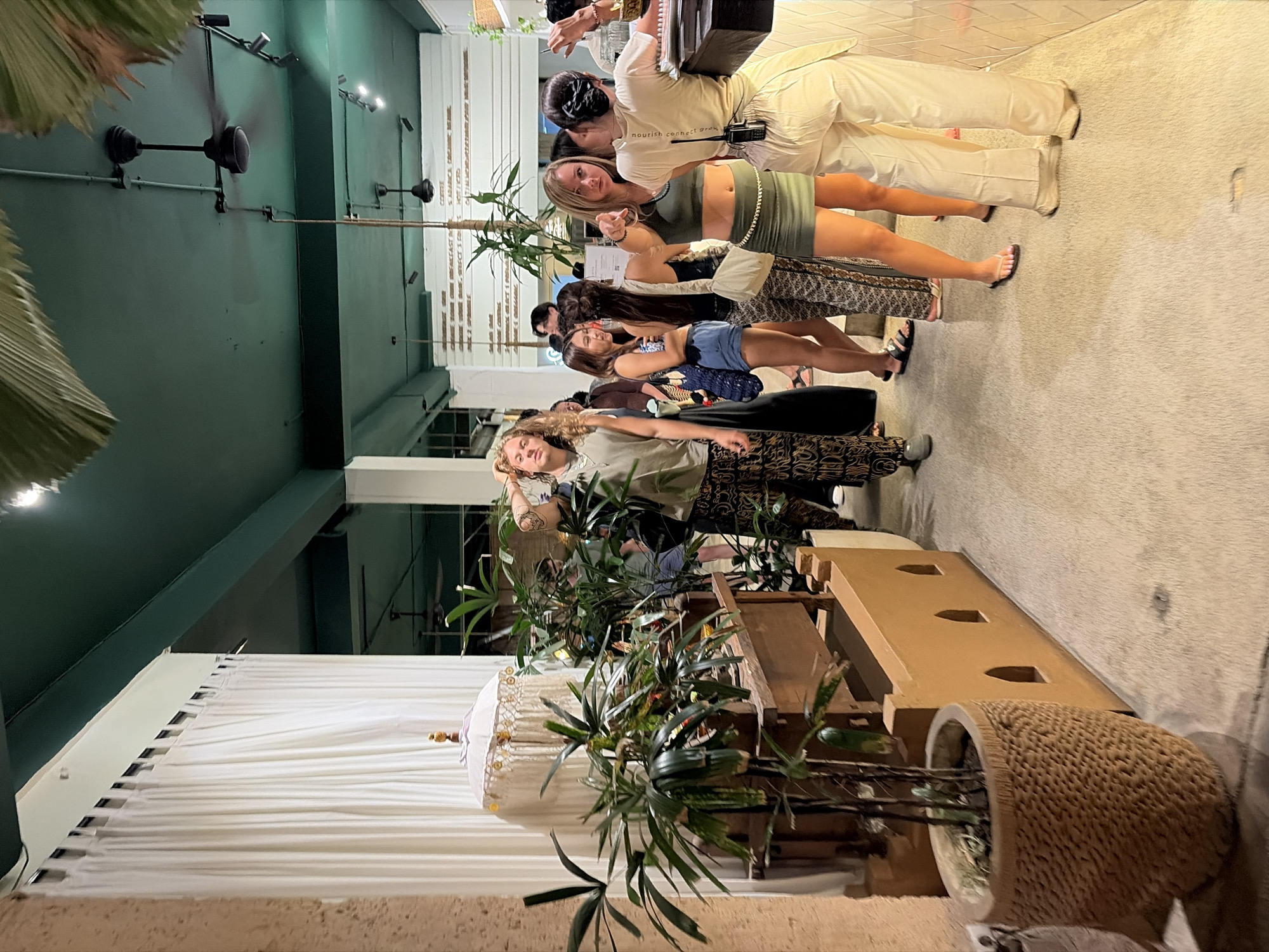 The image depicts a queue of people inside a dimly lit, open-air establishment in Ubud, Indonesia. The interior features a dark green ceiling with visible ventilation systems and black ceiling fans, contrasting with white pleated curtains along one wall and textured terracotta-colored walls.
In the foreground, a large, intricately woven brown planter holds a lush, leafy green plant. To the right, a low, terracotta-colored wall with arched openings runs along the edge of the walkway.
A group of individuals is visible, seemingly waiting in line. The person closest to the camera, a woman wearing a green crop top and a matching green skirt, is pointing towards the viewer. She has blonde hair and is accessorized with a chain belt. Next to her, a woman with dark hair in a bun wears a light-colored t-shirt and loose-fitting white pants, with a dark object clipped to her waistband.
Further back in the queue, a person with long, wavy blonde hair, wearing a light olive-colored sleeveless top and patterned pants, stands with their hand on their head. Other individuals in the line are dressed in casual attire, including shorts, patterned pants, and sleeveless tops, with some carrying bags.
The overall ambiance suggests a trendy, possibly cafe or boutique, setting, with natural elements like plants and earthy-toned structures contributing to the aesthetic. A menu board is partially visible on a white wall in the background. The lighting appears to be ambient, with spotlights illuminating certain areas.