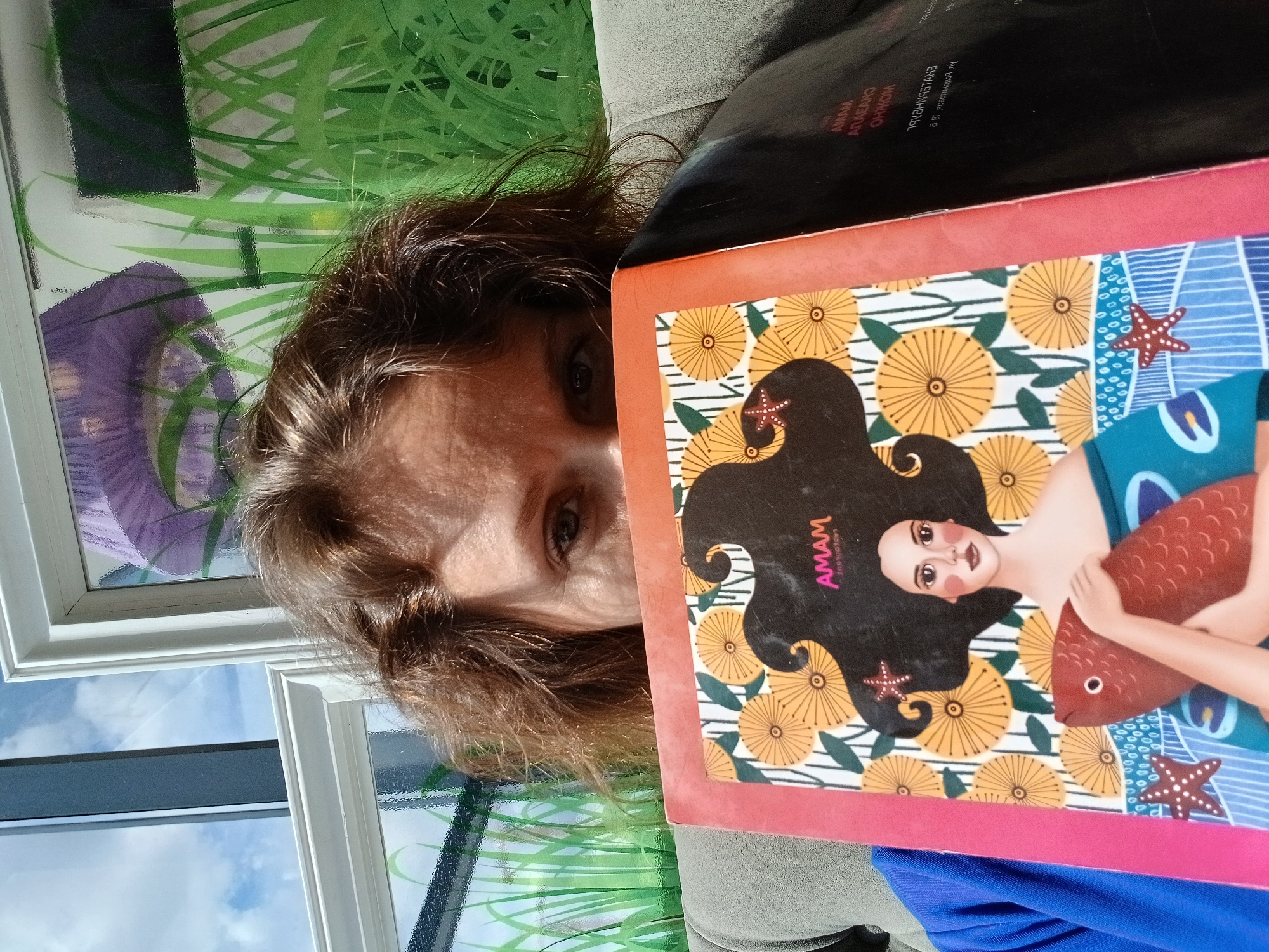 An adult with light brown hair is looking over a book with a colorful illustration of a woman holding a red fish. The woman is wearing a blue shirt. The book's cover features yellow flowers and starfish. The background is out of focus but appears to be an indoor setting with a window showing a blue sky and clouds, and a wall decorated with green grass and a purple object. Text is visible on the book cover and on a black surface to the right. The scene is likely in Yekaterinburg, Russia, based on the metadata.