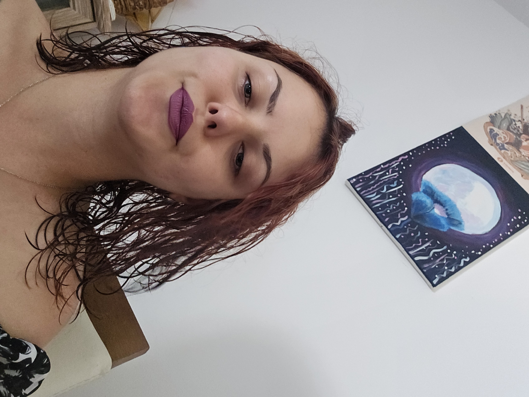 A medium shot captures a woman with red hair styled in a top knot and some strands loose and wet, framing her face. She is looking directly at the camera, her head slightly tilted. She is wearing purple lipstick and a silver necklace. Her shoulders and upper chest are visible. She is seated in front of a white wall where two framed artworks are displayed. The artwork on the left is partially visible and appears to depict figures. The artwork on the right is a painting of a large, round, blue object against a dark, starry background. To the left of the woman is a wooden nightstand with carved details. The environment appears to be indoors.