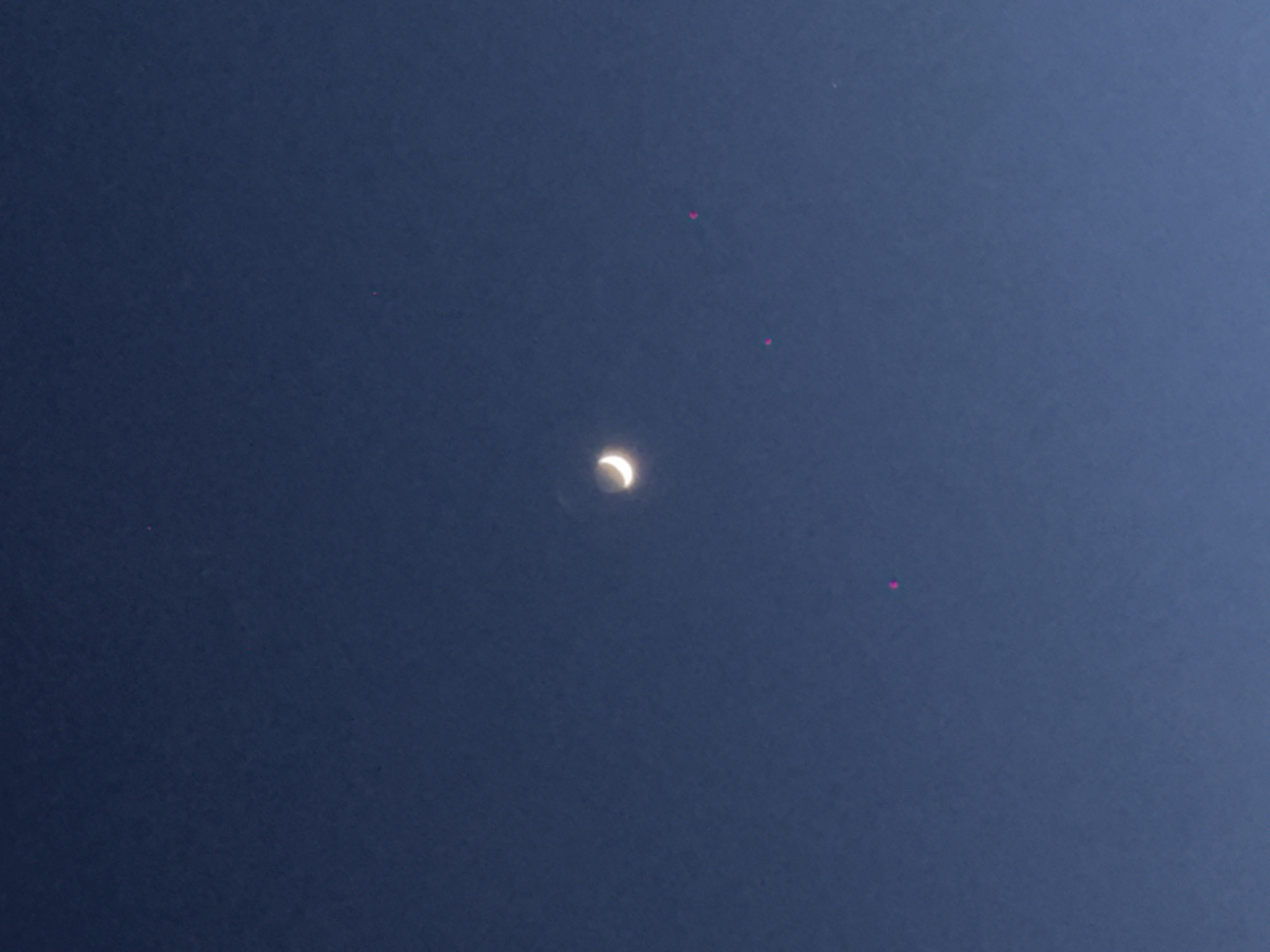 The image displays a celestial scene featuring a crescent moon against a deep, dark blue sky. The moon, positioned slightly above the center, is brightly illuminated in a crescent phase, with its unlit portion faintly discernible, suggesting either a partial lunar eclipse or earthshine. The sky is clear, devoid of clouds, indicating fair weather.
Scattered across the dark blue expanse are several small, bright, pinkish-red anomalies, appearing as distinct dots. Three prominent dots are visible: one in the upper left quadrant, another above the moon towards the upper center, and a third in the mid-right section of the sky. These dots are likely lens flares or sensor artifacts rather than celestial bodies.
The overall dark blue hue of the sky suggests the photograph was taken during twilight hours (dawn or dusk) or at night, allowing the moon to stand out prominently. The scene is set in Khimki, Russia. No people, other distinct objects, or visible text are present in the image.