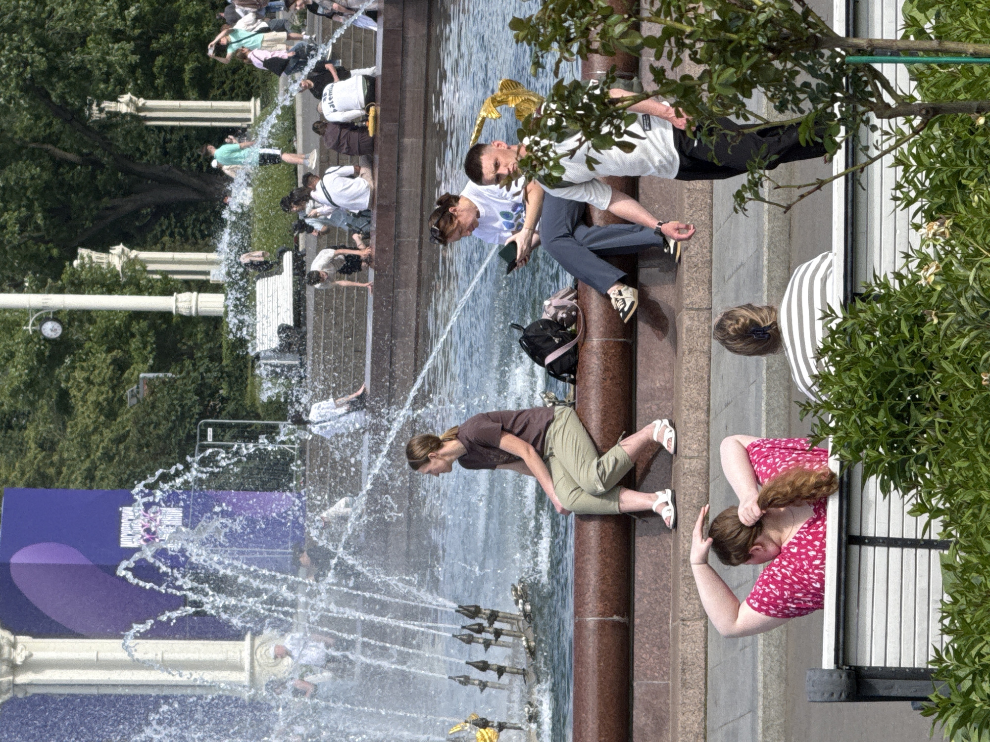 This image depicts a sunny day in Moscow, Russia, near a large fountain. The scene is filled with people enjoying the water and the atmosphere.
In the foreground, a woman in a pink and white patterned dress is seen adjusting her hair, with a bench and lush green foliage to her right. To her left, a woman in light green pants and a brown t-shirt is leaning forward, looking down at the fountain. She is wearing white sandals.
Further back, several other individuals are present. Some are sitting, while others are standing or walking near the fountain's edge. One man is wearing a light blue t-shirt and black shorts, while another is in a white t-shirt and blue pants. A group of people are gathered on stone steps behind the fountain, some appearing to be watching the water display.
The fountain itself is a prominent feature, with water spraying upwards in various streams, creating a dynamic and refreshing visual. In the background, trees and architectural elements suggest an urban park or public square. The overall impression is one of a lively and pleasant summer day. The vibrant purple banner on the left side of the image contains some indecipherable text.