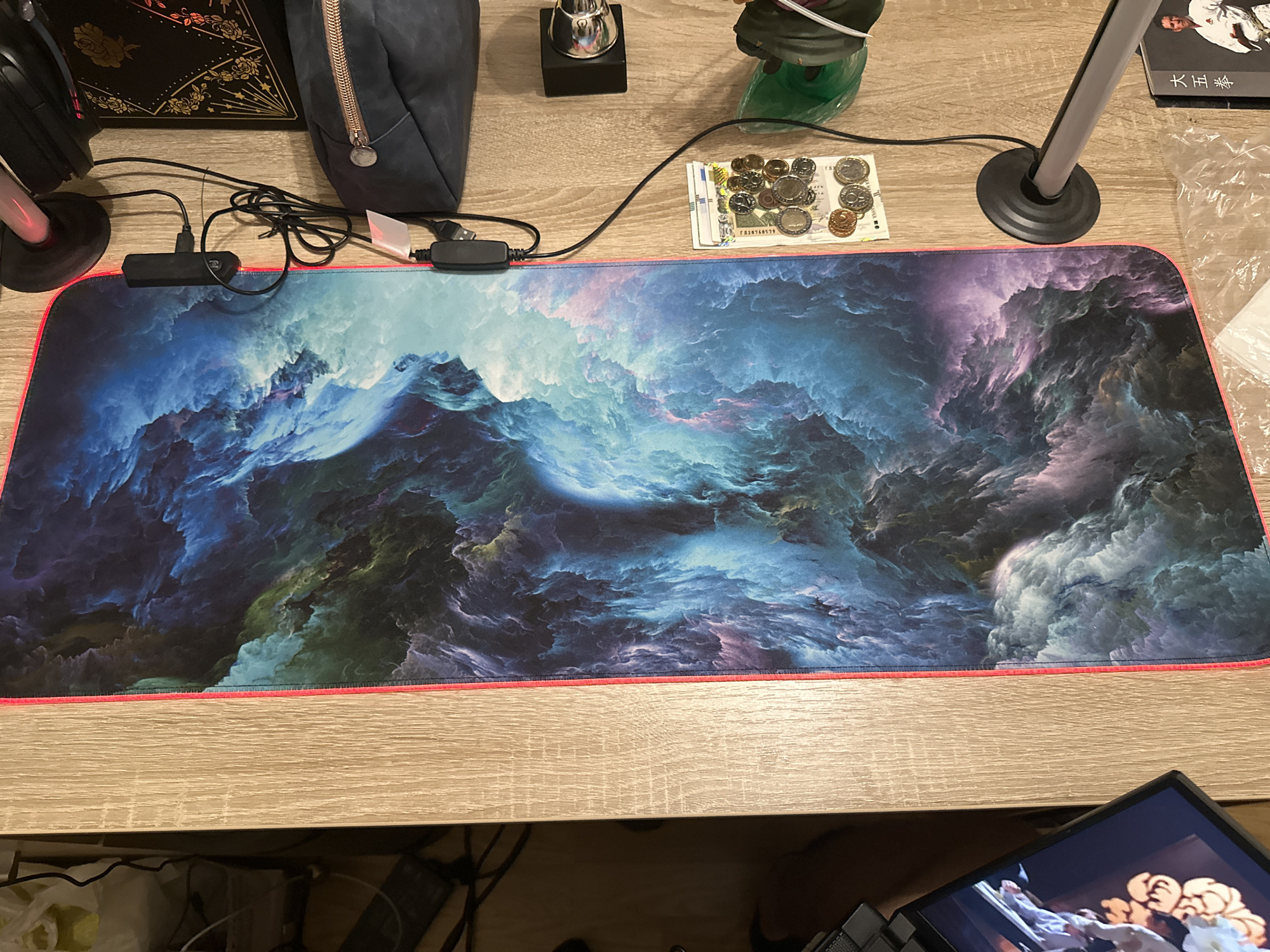 The image displays a top-down view of a desk with various items. A large, rectangular mousepad with a colorful, nebula-like pattern is the central focus. The mousepad has an RGB LED border that is illuminated with a pinkish-red hue. To the right of the mousepad, there is a stack of coins and some folded bills, suggesting a workspace. Above the coins and bills, there is a small black rectangular device connected to wires, likely part of the mousepad's lighting system. Further to the right, a dark blue pouch with a zipper and a black book with gold floral designs are visible. A black headset with a pink illuminated headband rests on a stand to the top right of the frame. A black object, possibly a charger, is also visible near the headset. In the lower right corner, a book with Chinese characters and a picture of a martial artist is partially visible. In the lower left corner, a laptop screen displays an image. The desk surface appears to be made of light-colored wood. The overall scene is one of a person's organized but eclectic workspace. The location is unknown.