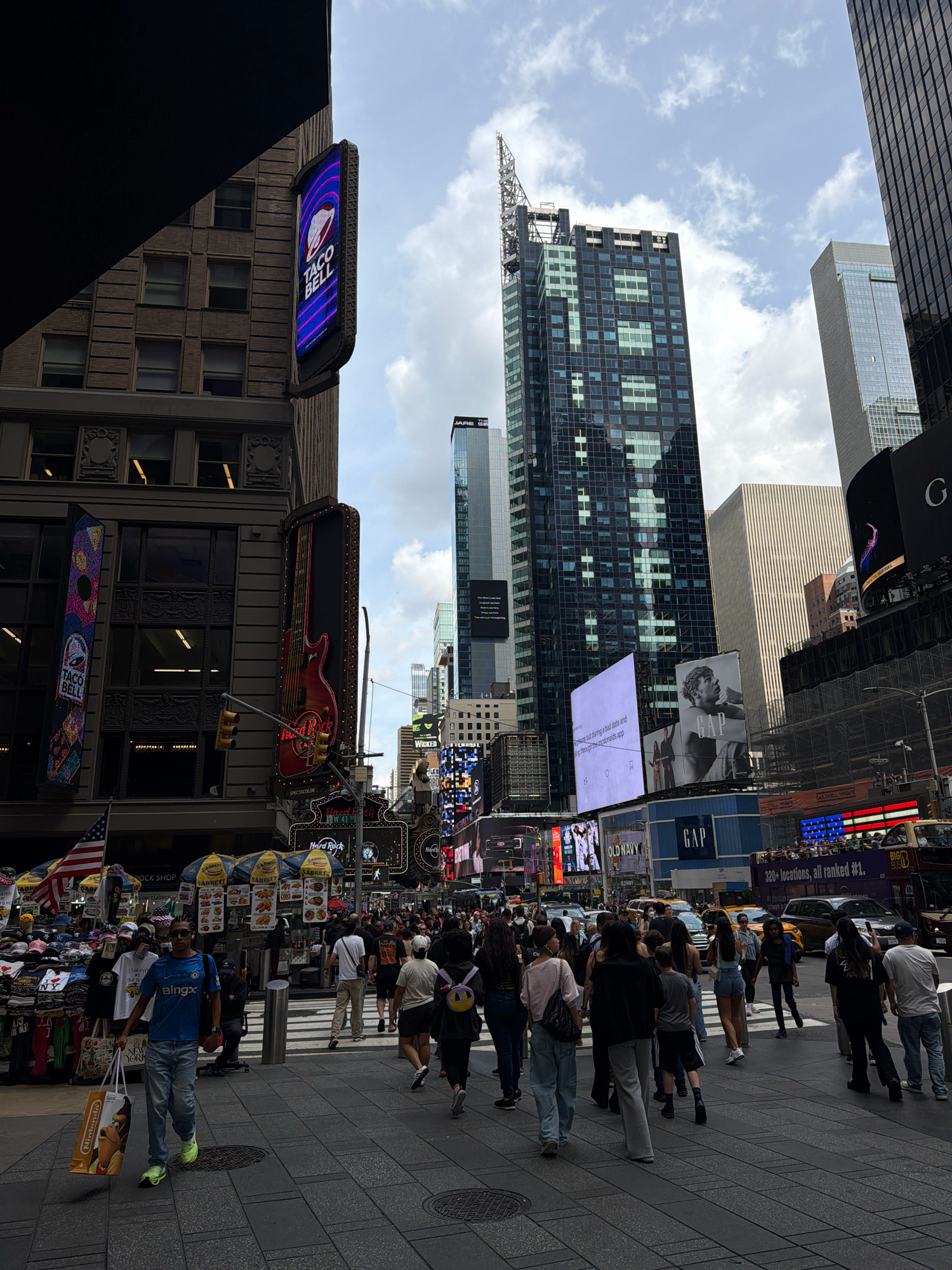 This is a medium-shot, eye-level, outdoor, daytime, full-color photograph taken in Times Square, New York City. The image captures a bustling street scene with a crowd of people walking on the sidewalk and crossing a street. Several large buildings with numerous windows and illuminated billboards dominate the background.
In the foreground, a man walks towards the viewer, carrying two shopping bags, one of which has a "Nintendo" logo. He is wearing a blue t-shirt, light-colored jeans, and bright green sneakers. Several other pedestrians are visible in the mid-ground, some walking in groups and others individually. A street vendor with multiple umbrellas is set up on the right side of the sidewalk, displaying various merchandise. Traffic, including cars and a double-decker bus, is present on the street in the background.
Notable signage includes a vertical "TACO BELL" sign on a building on the left, a large illuminated guitar shape with "Hard Rock" lettering above it, and various digital billboards displaying advertisements. The sky is partly cloudy. The overall impression is of a busy urban environment with a high volume of pedestrian and vehicle traffic.