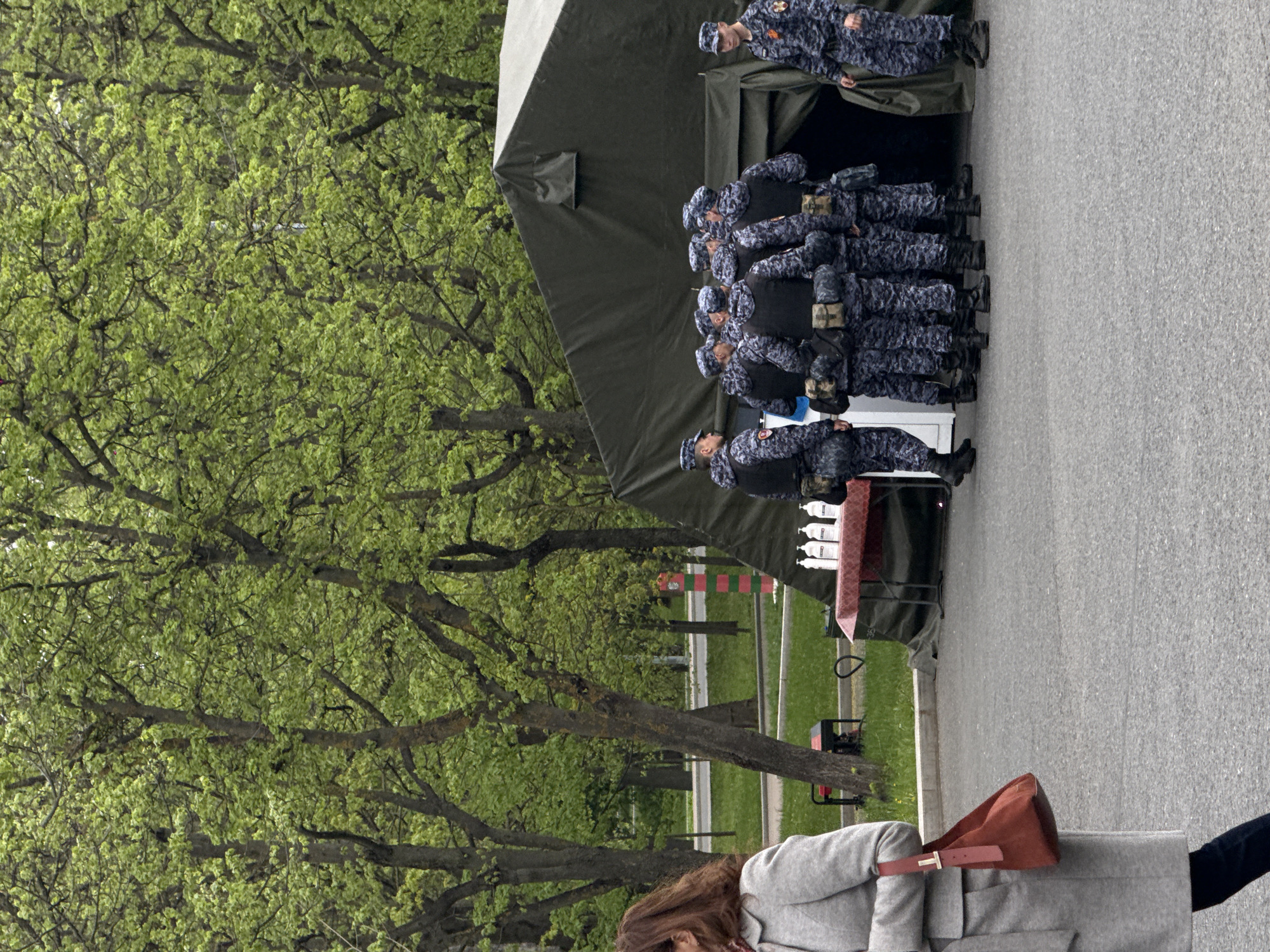 The image shows a group of people in matching blue camouflage uniforms standing near a large green tent in what appears to be a public space. The people are lined up closely together. A woman in a gray coat and brown purse is partially visible in the lower right corner of the image. Lush green trees are visible to the left of the tent. A road and grassy areas with benches and other structures are visible in the background. Based on the overall context and details such as the style of uniform, tent, and urban landscape, the location is Moscow, Russia.