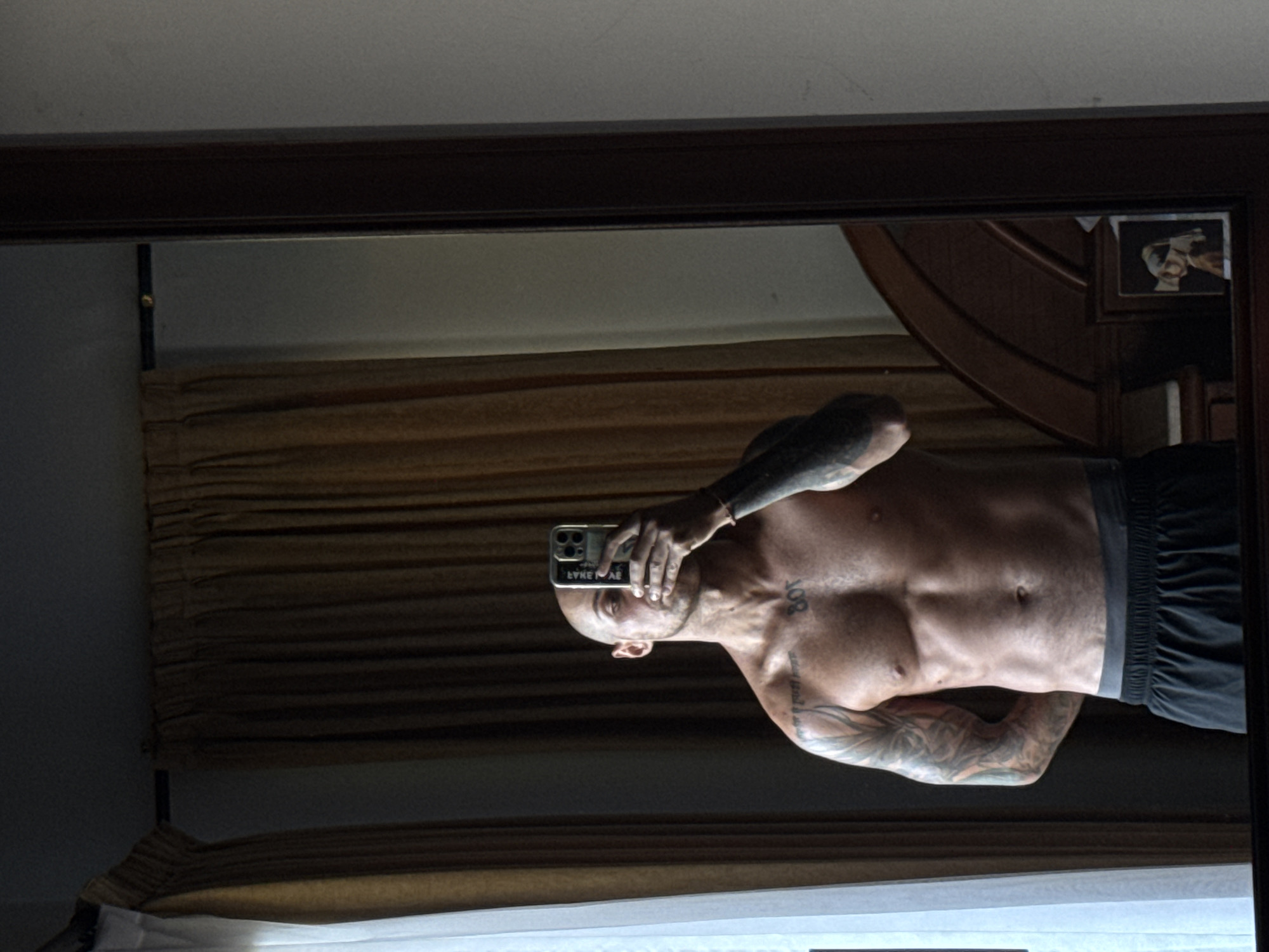 A shirtless, bald man with extensive tattoos on his arms and chest stands in front of a full-length mirror, taking a selfie with his phone. He is wearing dark shorts. Behind him, brown curtains are visible, and to his left, a white sheer curtain hangs. In the reflection of the mirror, a wooden headboard of a bed and a framed picture of "Girl with a Pearl Earring" are visible. The image appears to be taken indoors, likely in a bedroom, in Kuta Selatan, Indonesia.