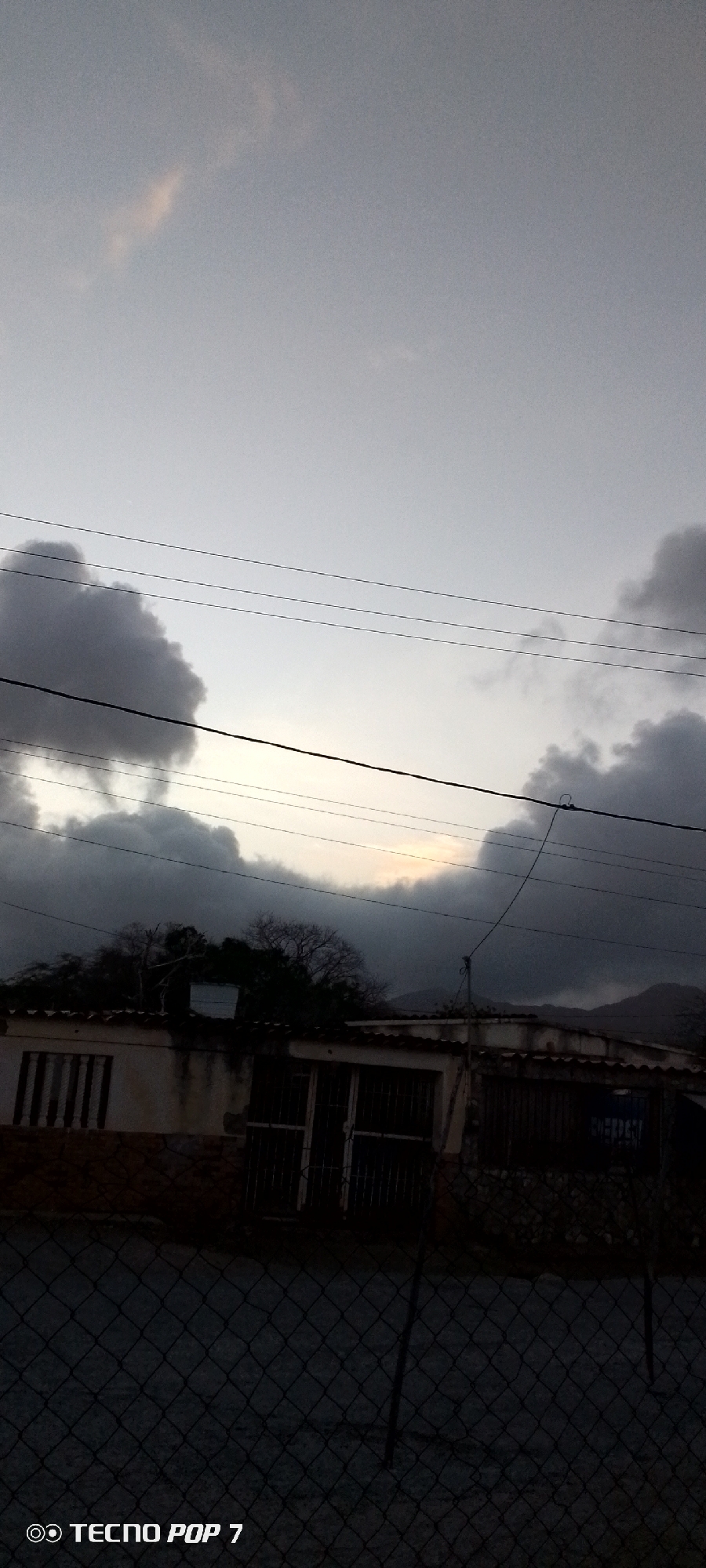 The photograph shows a view of a residential area in Altagracia, Venezuela, under a cloudy sky. A low building with a tile roof and barred windows and doors is visible behind a chain-link fence. Utility wires stretch across the sky. Trees are visible behind the building. In the distance are mountains. The image was taken with a Tecno Pop 7.
