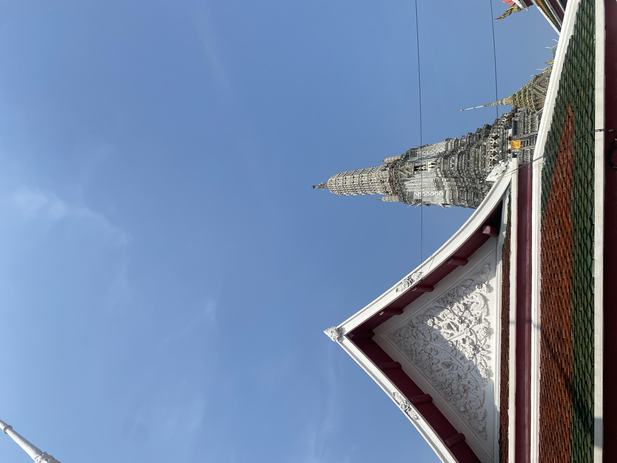 The image shows a low-angle view of Wat Arun Ratchawararam Ratchawaramahawihan, commonly known as Wat Arun or the Temple of Dawn, in Bangkok, Thailand. The image is taken from below, looking upwards towards the temple's main prang (tower) against a clear, blue sky. In the foreground, a portion of the temple's roof is visible, featuring intricately carved white ornamentation and tiles in reddish-brown and green hues.