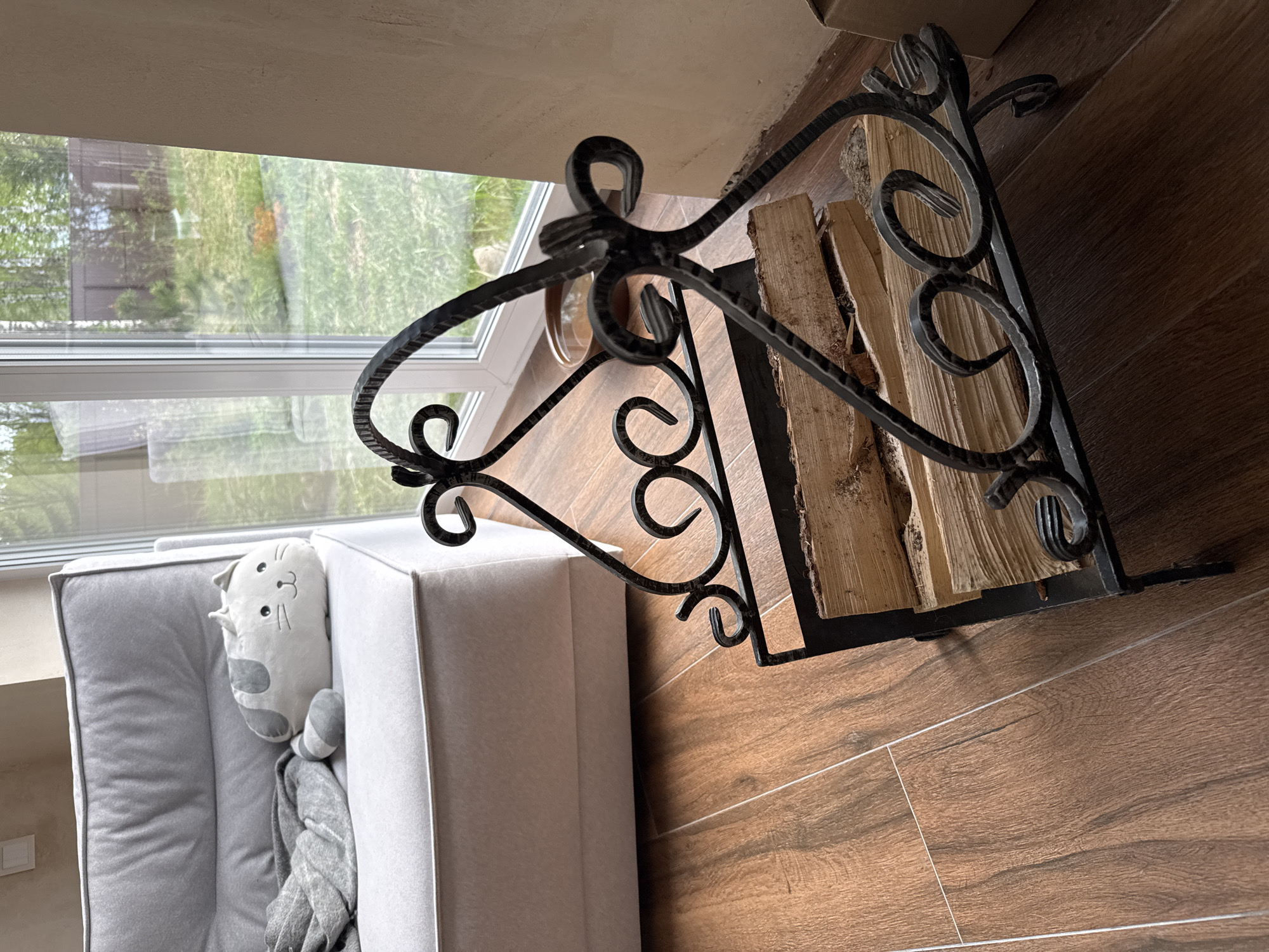 The image presents an interior scene. Dominating the view is a black, ornate wrought-iron log holder containing several pieces of wood. To the left is a gray sectional sofa with a stuffed cat toy positioned on the corner section of the sofa. A gray blanket is draped over the side of the sofa. Behind the sofa is a window offering a view of an exterior space featuring green grass. The floor is made of brown wooden planks. There is no evidence to determine if this is the city of Steklyannyi, Russia. No people are visible in the image.