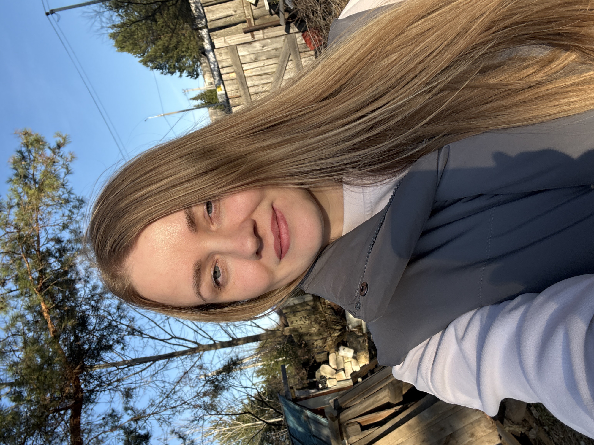 A close-up outdoor portrait features a young woman with long, straight, blonde hair. She is looking directly at the camera with a gentle, relaxed smile, her head slightly tilted. She has light-colored eyes and her lips are subtly colored with pink lipstick. She is dressed in a dark grey puffer vest worn over a light blue or off-white long-sleeved top.
The background is bathed in bright, natural sunlight under a clear blue sky. To the left, a tall, green pine tree is visible, while other trees, some deciduous with bare branches, frame the scene. Behind and to the right of the woman, various weathered wooden structures, resembling a fence or stacked planks, are present, alongside scattered light-colored bricks or stones. Thin power lines are faintly visible stretching across the sky. The strong sunlight creates highlights on her hair and face, and casts subtle shadows, indicating a sunny daytime setting. The scene evokes a rustic or suburban outdoor environment, consistent with a location like Chaikovsky, Russia. There is no visible text in the image.