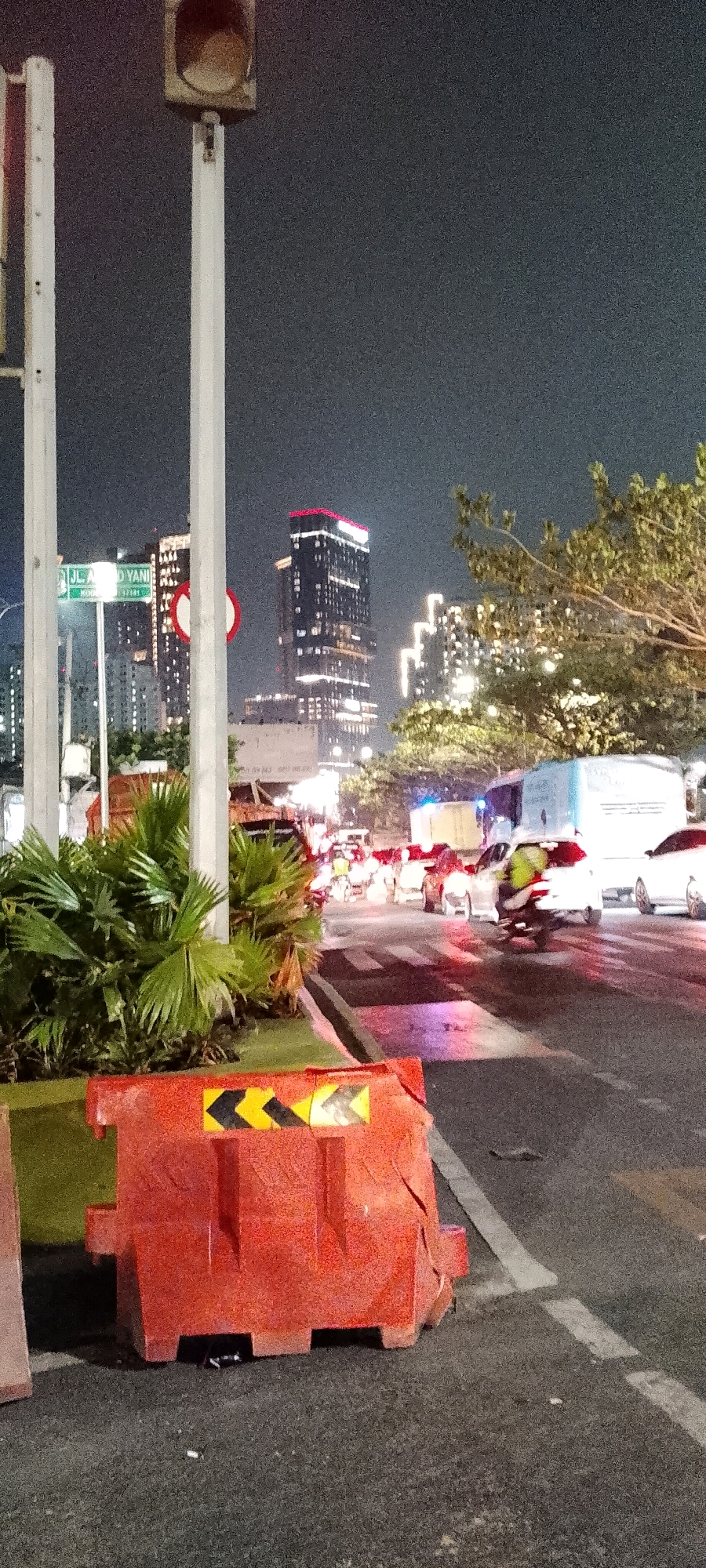 The image depicts a nighttime street scene in Bekasi, Indonesia, likely on Jalan Ahmad Yani, a major thoroughfare. A traffic jam is visible, with numerous cars and motorcycles moving slowly. A white truck is prominent in the middle ground. In the foreground, a bright orange traffic barrier with yellow and black directional arrows is placed on the asphalt road. Behind the barrier, lush green palm-like plants provide a touch of nature amidst the urban setting. In the background, towering skyscrapers, illuminated with lights, dominate the skyline, indicative of a bustling city. The overall atmosphere is one of nighttime urban activity, with the traffic jam suggesting a busy period.