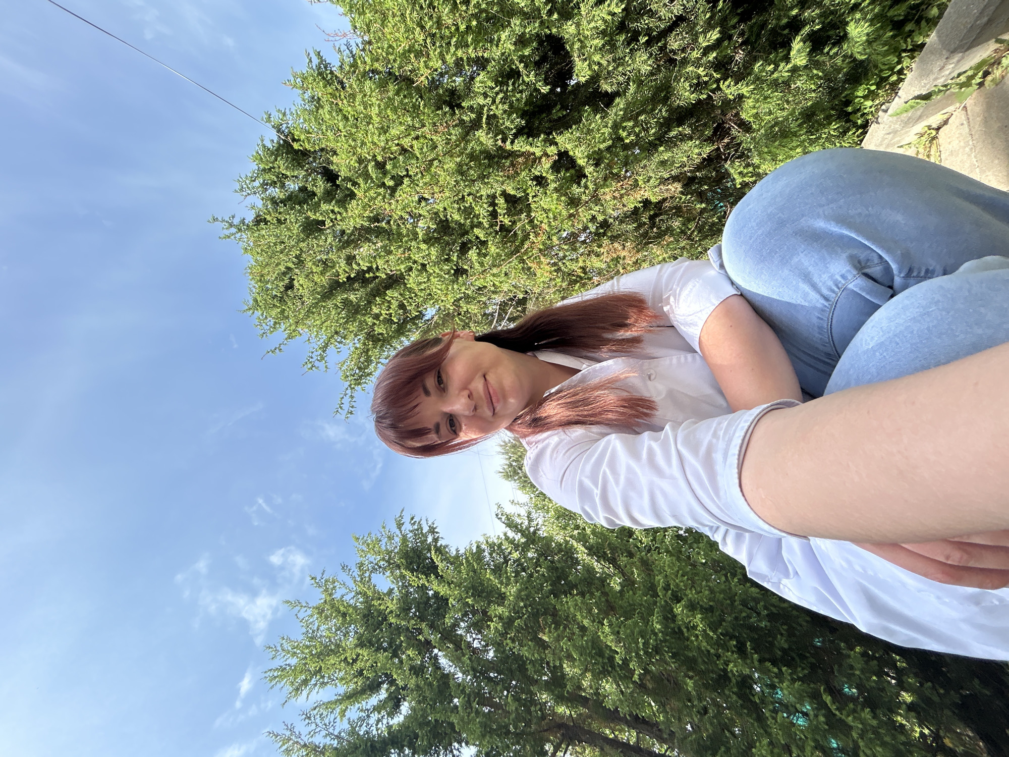 A selfie taken from a low angle shows a young woman in Фоки, Russia, seemingly sitting outside. Her face is slightly off-center, framed by auburn hair with bangs. She wears a white shirt and denim jeans. Behind her, two large, green trees reach upwards towards a partly cloudy blue sky. The trees appear to be located near the street, suggesting a park or green space within the city.