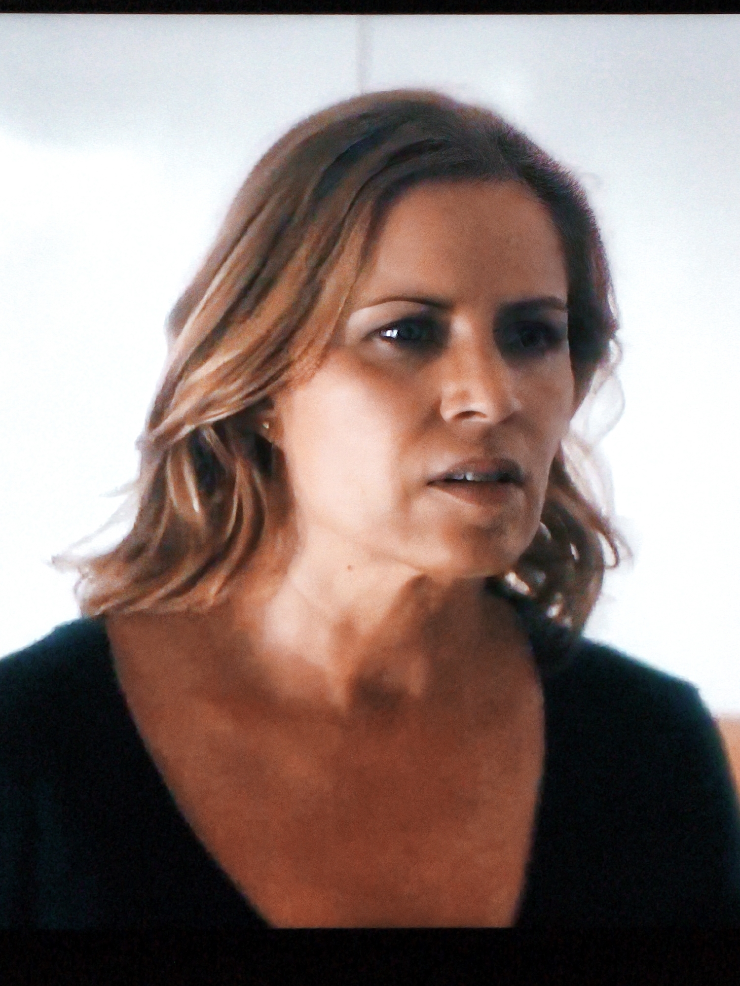 A medium shot, captured on a screen, shows a woman with shoulder-length, reddish-brown hair styled with loose waves. She is wearing a black top with a V-neckline. Her face is turned slightly to the right, and she appears to be looking off-camera. Her expression is serious, with her mouth slightly open. The background is a bright, out-of-focus white. The image quality is grainy, suggesting it was taken from a television or computer screen. The scene does not contain any identifiable landmarks or elements that would indicate a specific location in Vladivostok, Russia.
