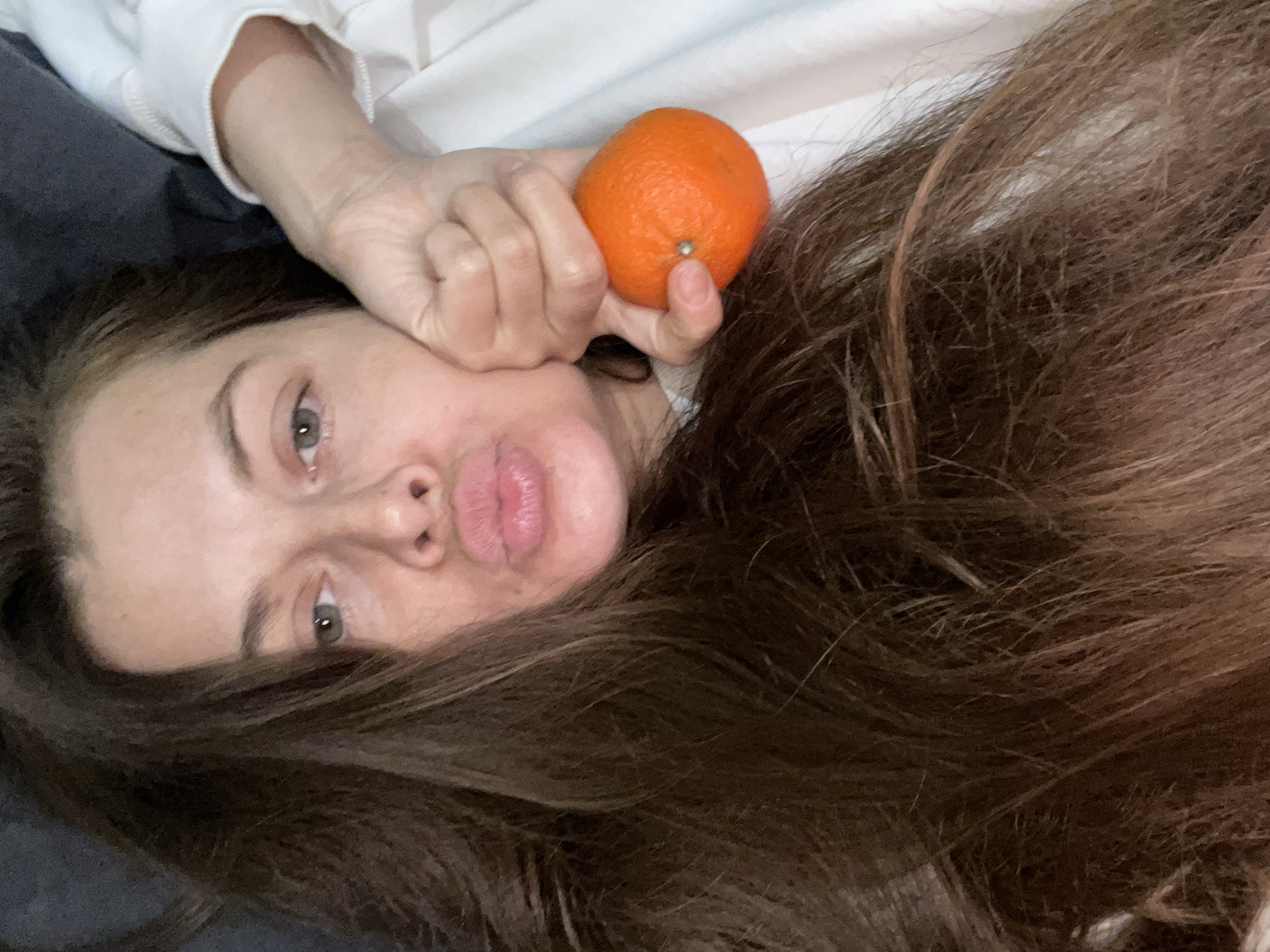 In a close-up, low-angle shot, a woman with long, dark brown hair lies on her side on what appears to be a bed, possibly in Заречный, Russia. Her hair is spread out around her face and shoulders. She is making a kissing face, with her lips puckered. Her right hand is raised, with her fist gently pressed against her cheek. In her hand, she holds a single, bright orange tangerine. The background is a soft, out-of-focus blend of dark gray and white, suggesting bedding or blankets. The lighting is soft and diffused, creating a natural, unposed feel.