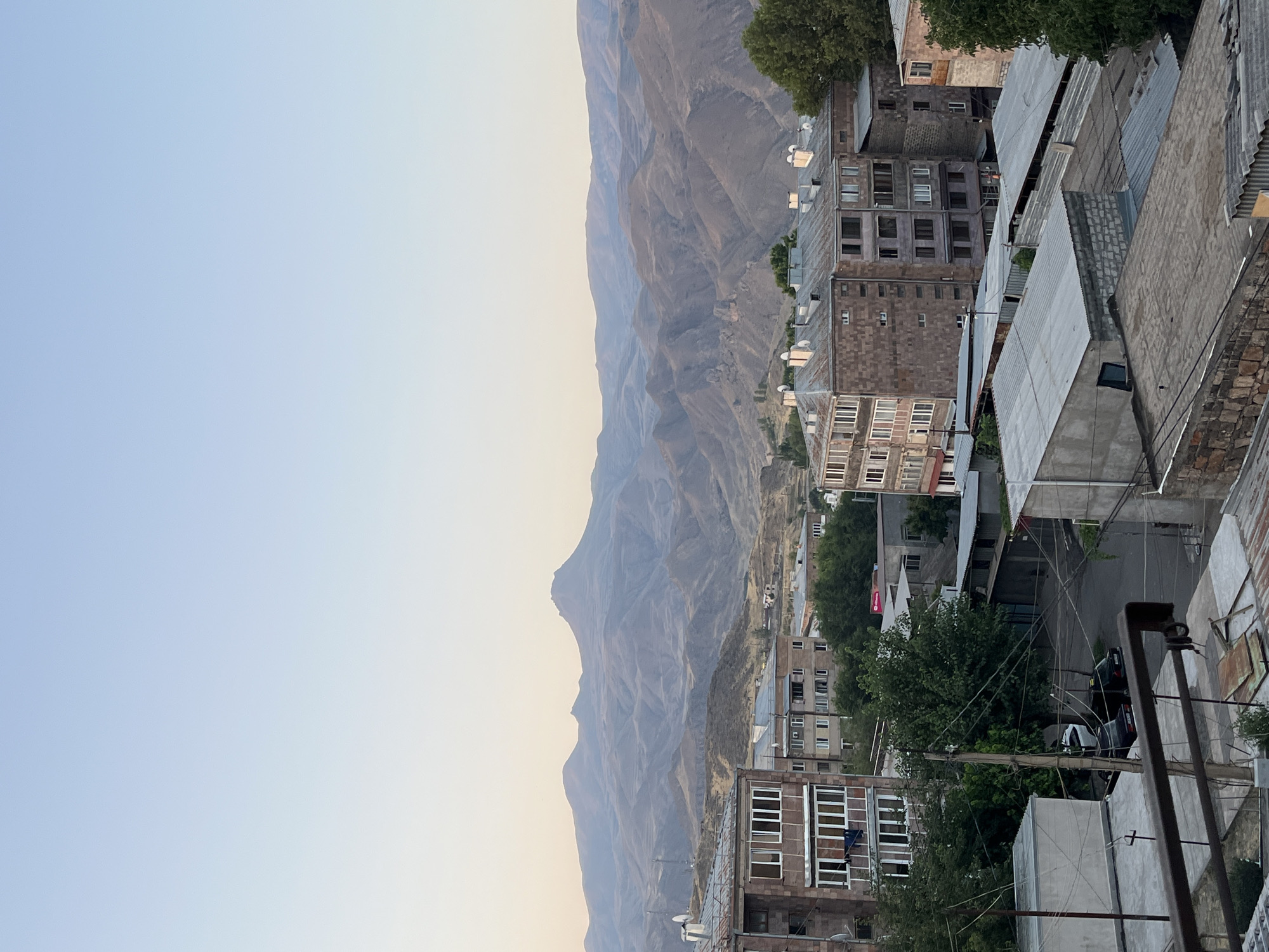 A high-angle, eye-level medium shot captures a view of the town of Gladzor, Armenia, with mountains in the background. The sky is clear and pale blue, transitioning to a lighter hue near the horizon. In the foreground, several buildings with flat roofs and balconies are visible. The architecture appears to be from the Soviet era, with multi-story apartment blocks constructed of brick and concrete. Some buildings have numerous windows and satellite dishes. Trees are interspersed among the buildings, with some appearing to be quite mature. A narrow street runs between the buildings, with two cars parked on its side. The middle ground is dominated by rolling hills and mountains, their slopes appearing dry and sparsely vegetated. The overall impression is one of a somewhat aged but functional urban environment nestled within a rugged natural landscape.