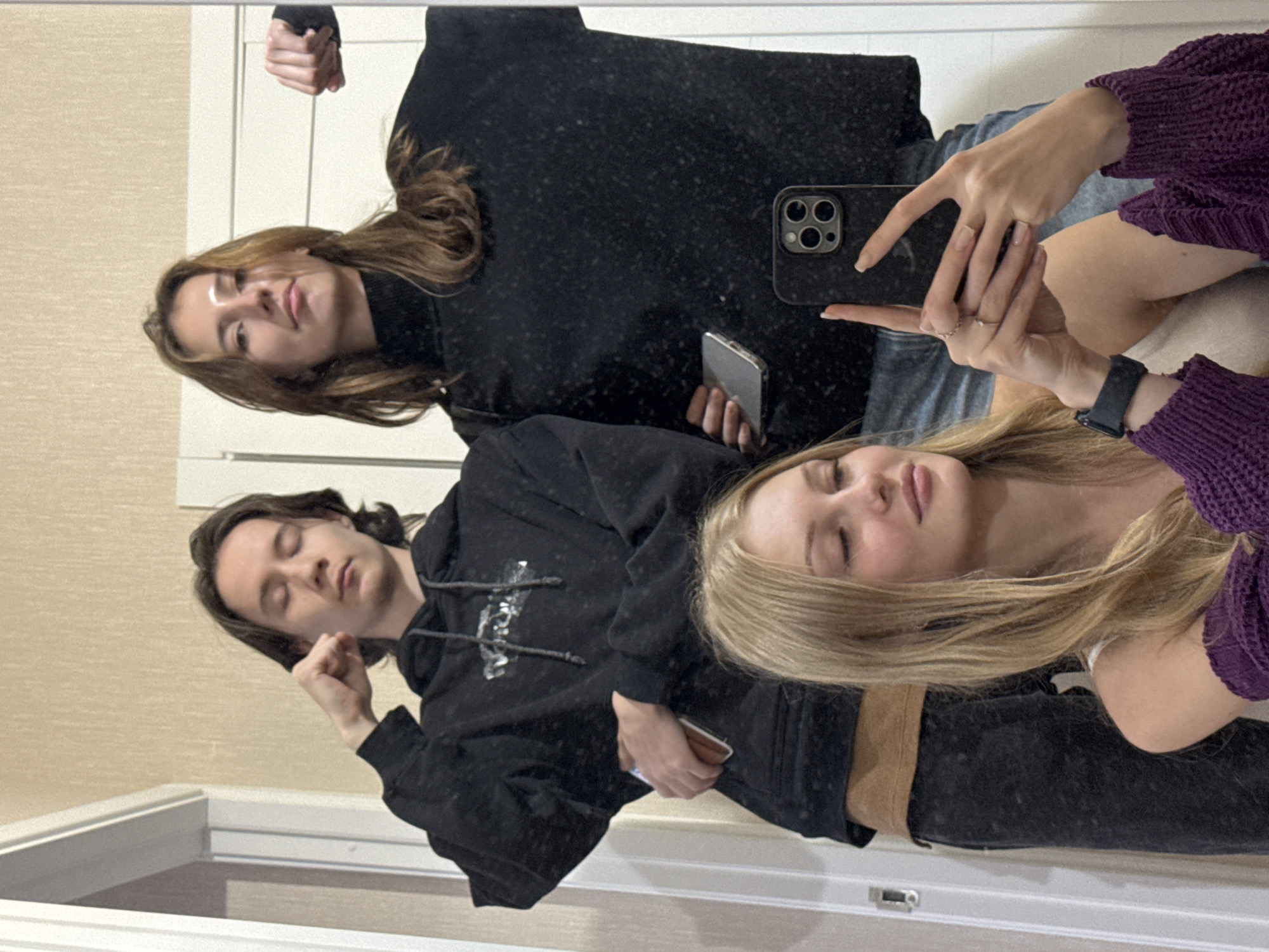 Three young adults are taking a selfie in a bathroom, reflected in a mirror. A woman with blonde hair is in the foreground, holding a smartphone and wearing a purple knitted sweater. Behind her, two individuals, one with long dark hair and the other with long brown hair, are posing, flexing their biceps. The background appears to be a typical bathroom with tiled walls and a door. The image does not provide any specific clues to pinpoint the location within the city of Perm, Russia.