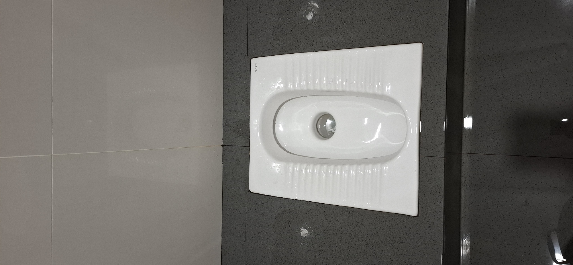The image shows a squat toilet with the brand name "TOTO" visible on its rim. The toilet is white and made of porcelain. It is positioned on a dark gray speckled floor, which is tiled. Behind the toilet, there are light gray tiles on the wall, separated by thin white grout lines. The environment appears to be a restroom. There are no people visible in the image. The image does not provide any specific location information that would allow for pinpointing it in Karangpandan, Indonesia.