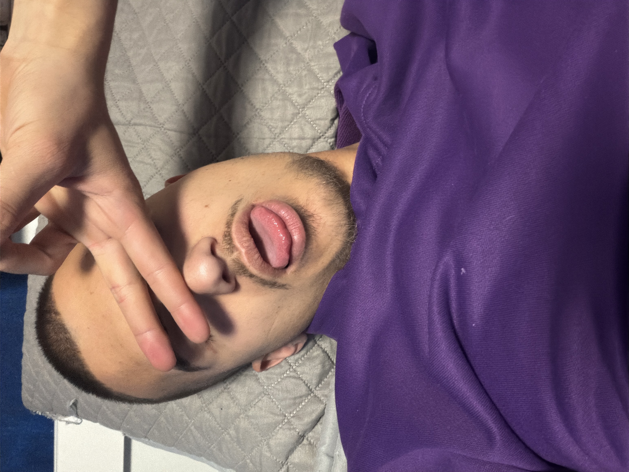 A person is lying on their back on a gray quilted surface, possibly a bed or a cushion. The person wears a purple shirt. The person's face is visible, with the mouth open and tongue protruding. A hand with fingers partially covering the face. The environment appears to be an indoor setting. There is no information about Sofia, Bulgaria in the image.