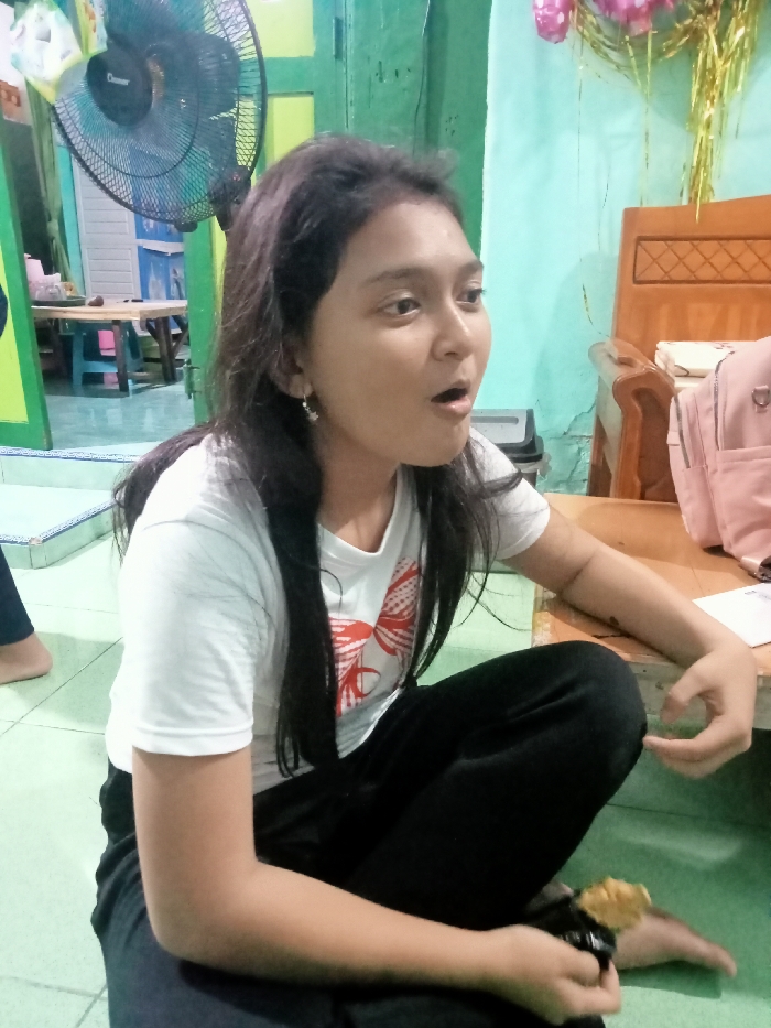 The image depicts an indoor residential or common area located in an unknown city. A female individual, appearing young, is seated on a light green floor mat with a blue and white patterned border, facing towards the right with her mouth open. She has long dark hair, wears a white short-sleeved t-shirt with a red graphic design on the chest, and black pants. Small silver-colored earrings are visible. Her left hand holds a dark object topped with a light-colored snack.
The background features a two-tone wall, light green on the left and light blue/teal on the right. A black stand fan is positioned on the left, near a doorway with a bright green frame leading to an adjacent room containing a light-colored table and assorted seating. To the right, a partial view of a wooden chair is visible, with pink and gold decorative elements suspended above it. A low, light-colored wooden surface next to the subject holds a pink backpack and a white envelope.