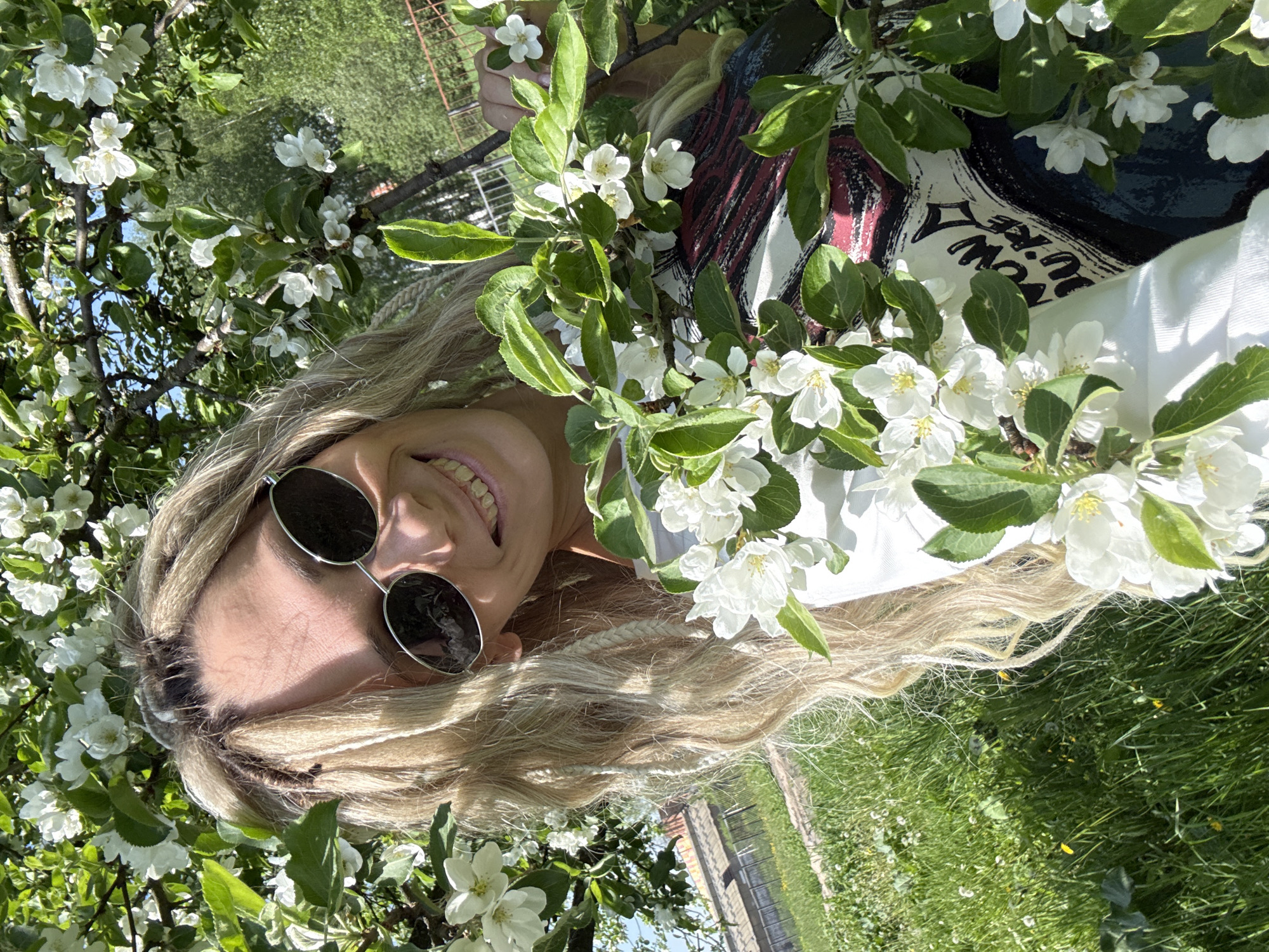 A woman with long blonde hair and round sunglasses poses amidst blooming apple blossoms in Rusanovo, Russia. She is smiling and wearing a white t-shirt with black text. The environment is a green, grassy area with a low fence in the background. The lighting suggests a sunny day.