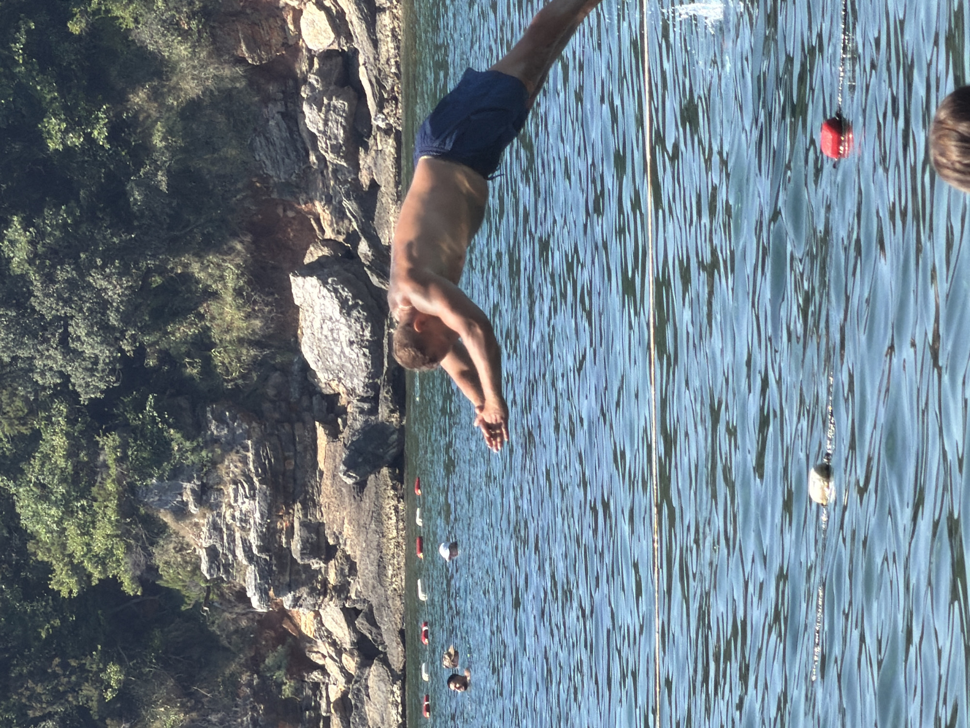 A man is captured mid-dive into the clear blue water of the Adriatic Sea, with rocky shores and lush green vegetation forming the backdrop. The man, wearing dark blue swim trunks, is horizontally positioned, his body arched as he plunges into the water. His arms are extended forward, preparing to enter the water. In the background, other swimmers are visible, some with their heads above the surface. The water is marked by a floating rope with buoys, and a bright red buoy is visible in the foreground. This scene is set in Budva, Montenegro, a popular coastal city known for its beautiful beaches and clear waters. The sun appears to be shining brightly, casting reflections on the water's surface.