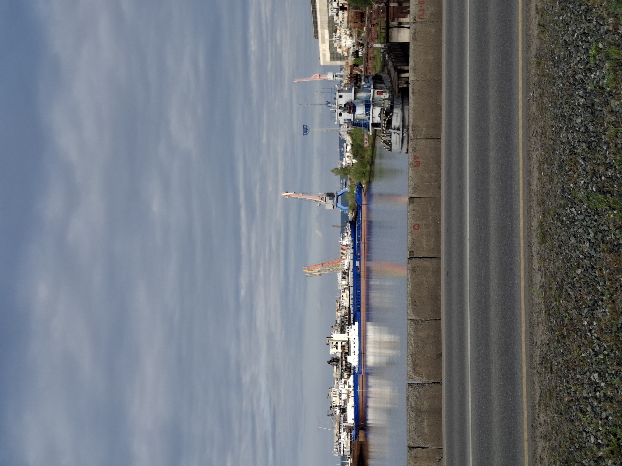 The image depicts a cloudy day at a shipyard in Chaikovsky, Russia. In the foreground, a paved road runs horizontally across the lower portion of the frame, bordered by a gravel and grassy shoulder. Behind the road, a concrete barrier wall separates it from the water. The water, appearing calm, reflects the sky and the vessels docked in the harbor.
The harbor is filled with numerous ships of various sizes, some appearing weathered or under repair. Prominent among them are several large barges, one of which is a blue barge with a white deck. A smaller, white tugboat is moored near a dock on the right side of the frame. Several large cranes, including a tall pink and orange one, are visible throughout the shipyard, suggesting ongoing operations. In the background, industrial buildings and more docked vessels can be seen. The overall scene conveys a working port or shipyard environment under a gray, overcast sky.