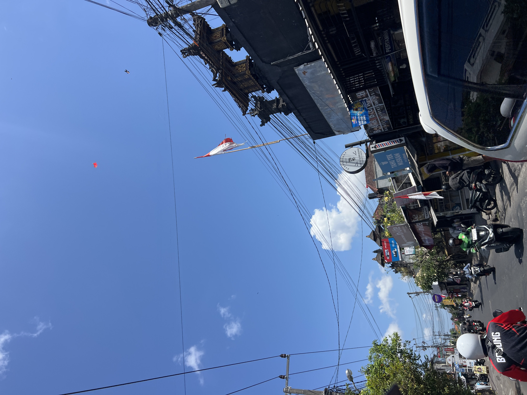 The image shows a street scene in Denpasar, Indonesia, on a clear, sunny day. The sky is bright blue with scattered white clouds. Overhead, a dense network of electrical wires crisscrosses the frame. In the distance, a red kite is visible against the sky.
The street is lined with buildings, many of which appear to be shops or businesses, indicated by signage. Some buildings have traditional architectural elements, such as ornate roofs. An Indonesian flag, red and white, is prominently displayed on a pole in front of one of the buildings.
The street is populated with various forms of transportation. Several motorcycles are being ridden by people wearing helmets. One person in the foreground is riding a motorcycle with a white helmet and a red and black jacket. Further down the street, more motorcycles and a white car are visible. People are also seen standing on the sidewalks. The overall impression is a busy, everyday street scene in an Indonesian city.